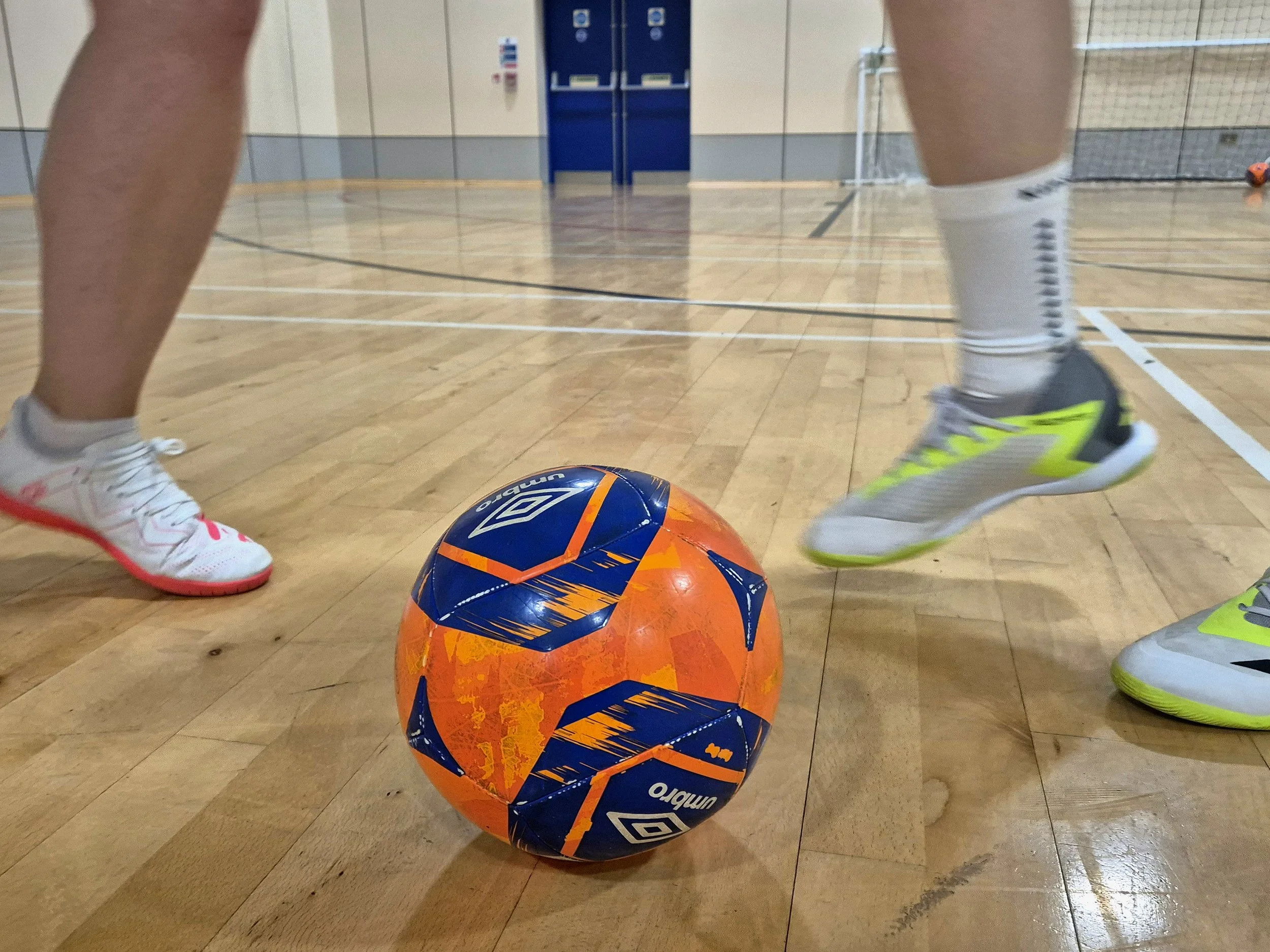 Two people in athletic shoes playing indoor soccer, with an orange and blue soccer ball on a wooden gym floor and a netted goal in the background.