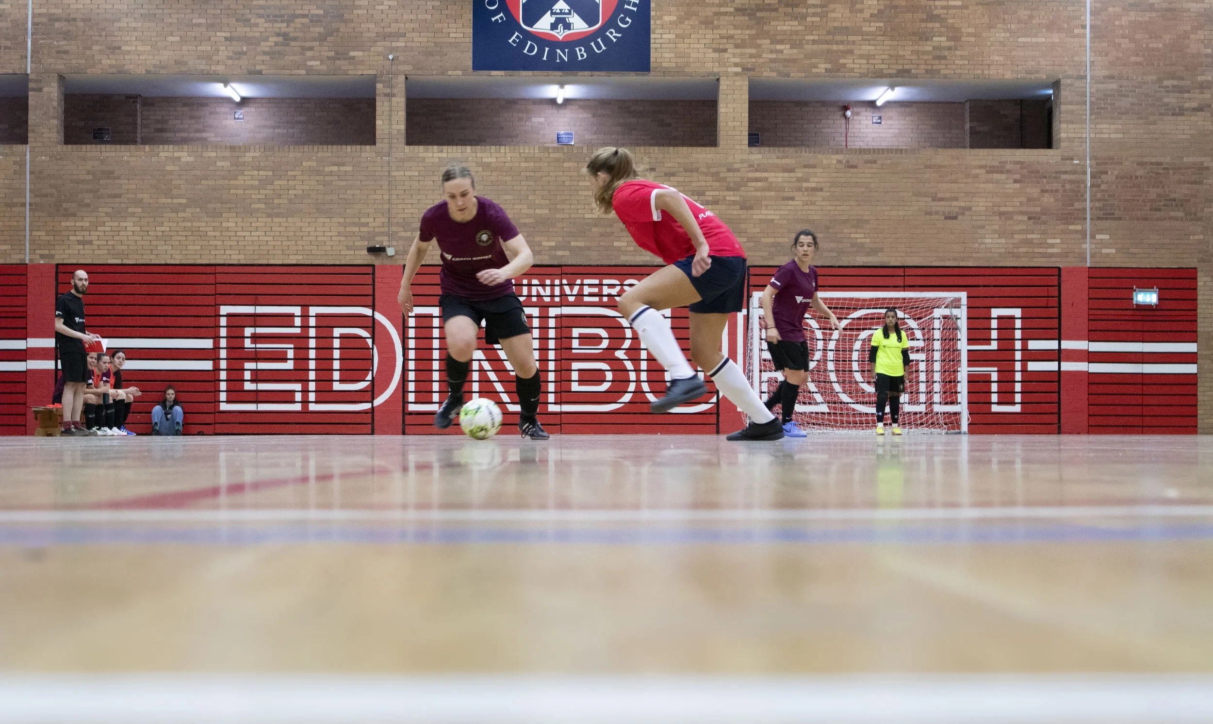 Two women playing futsal in an indoor gym, with one woman in a red jersey and the other in a purple jersey, trying to gain control of the soccer ball. A referee and three women sitting on the sidelines are visible in the background. The gym has a brick wall background with a banner and a small goal.