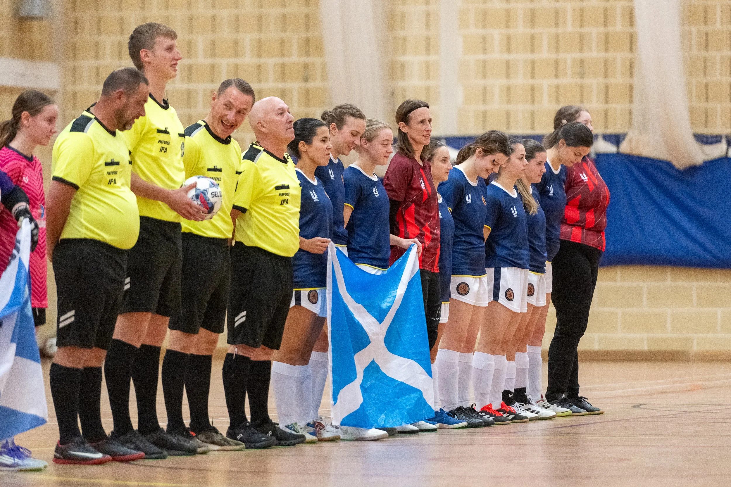 A group of female soccer players and referees standing in a line indoors, with some holding flags and a soccer ball, in a sports hall with a wooden wall background.