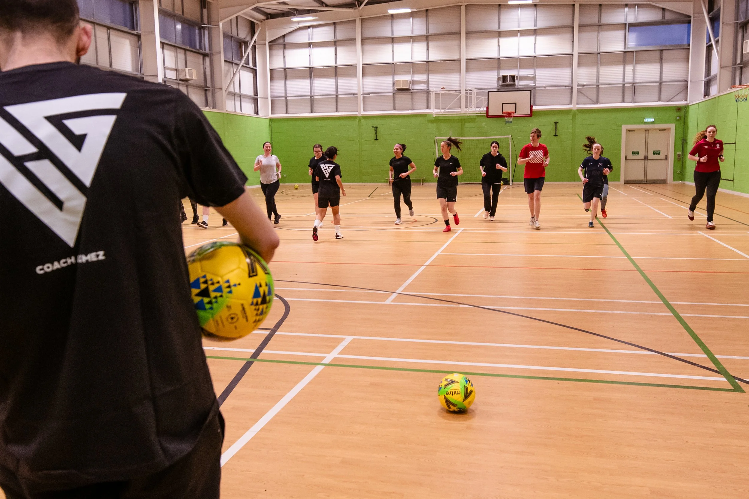 A group of women running inside a gymnasium, with a coach holding a yellow and blue soccer ball in the foreground. The gym has green walls and a basketball hoop.