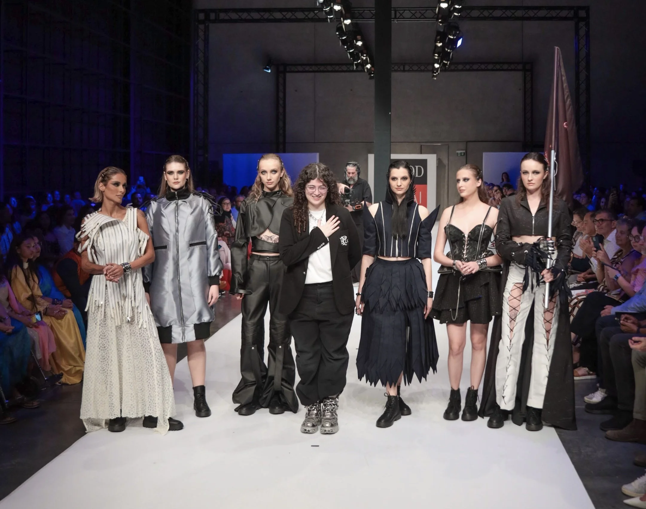 Models walking on runway at a fashion show with audience seated on both sides in a dark interior space.