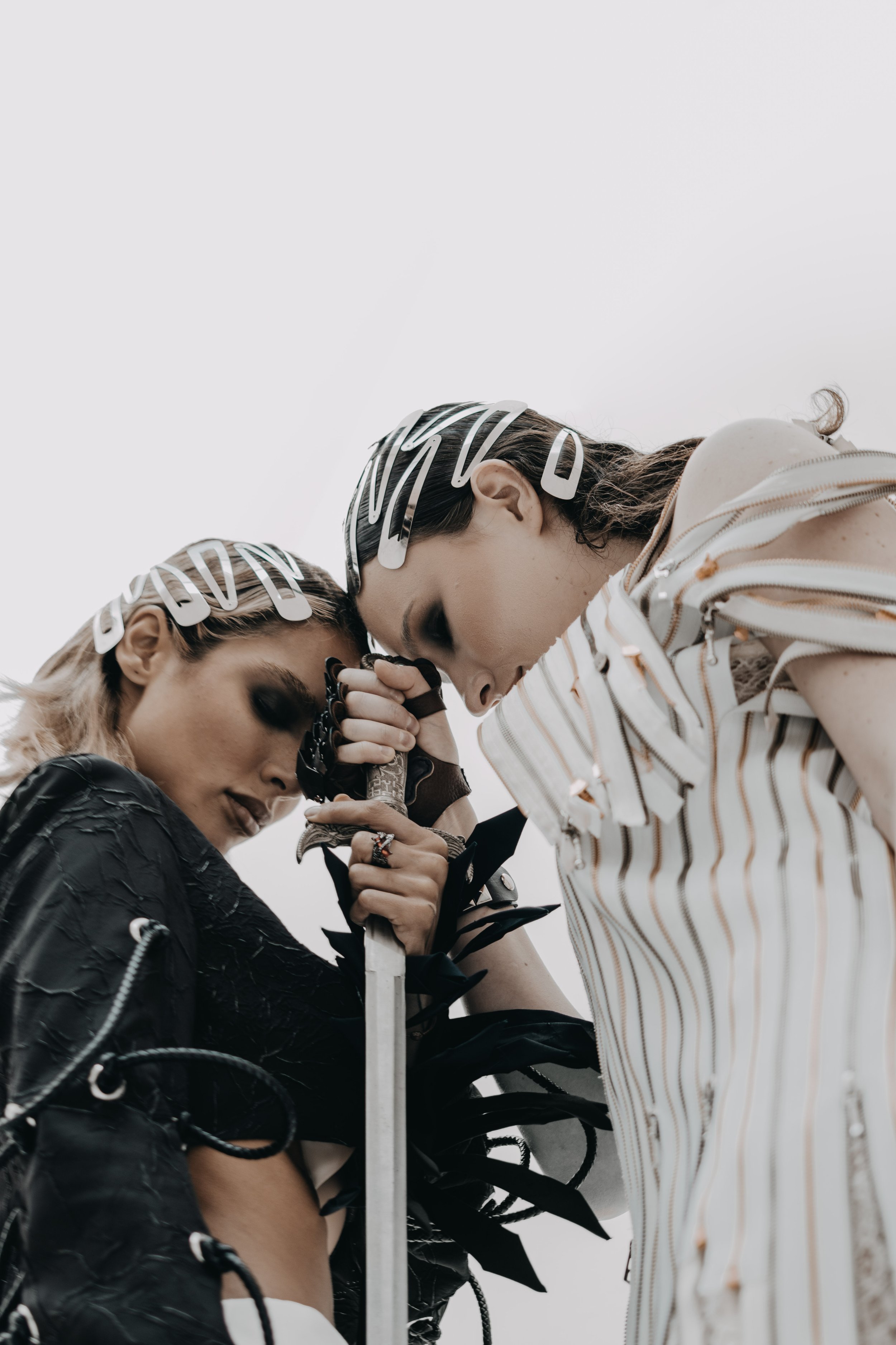Two women with futuristic accessories and dramatic makeup pose closely together, holding a sword between them, against a plain sky background.