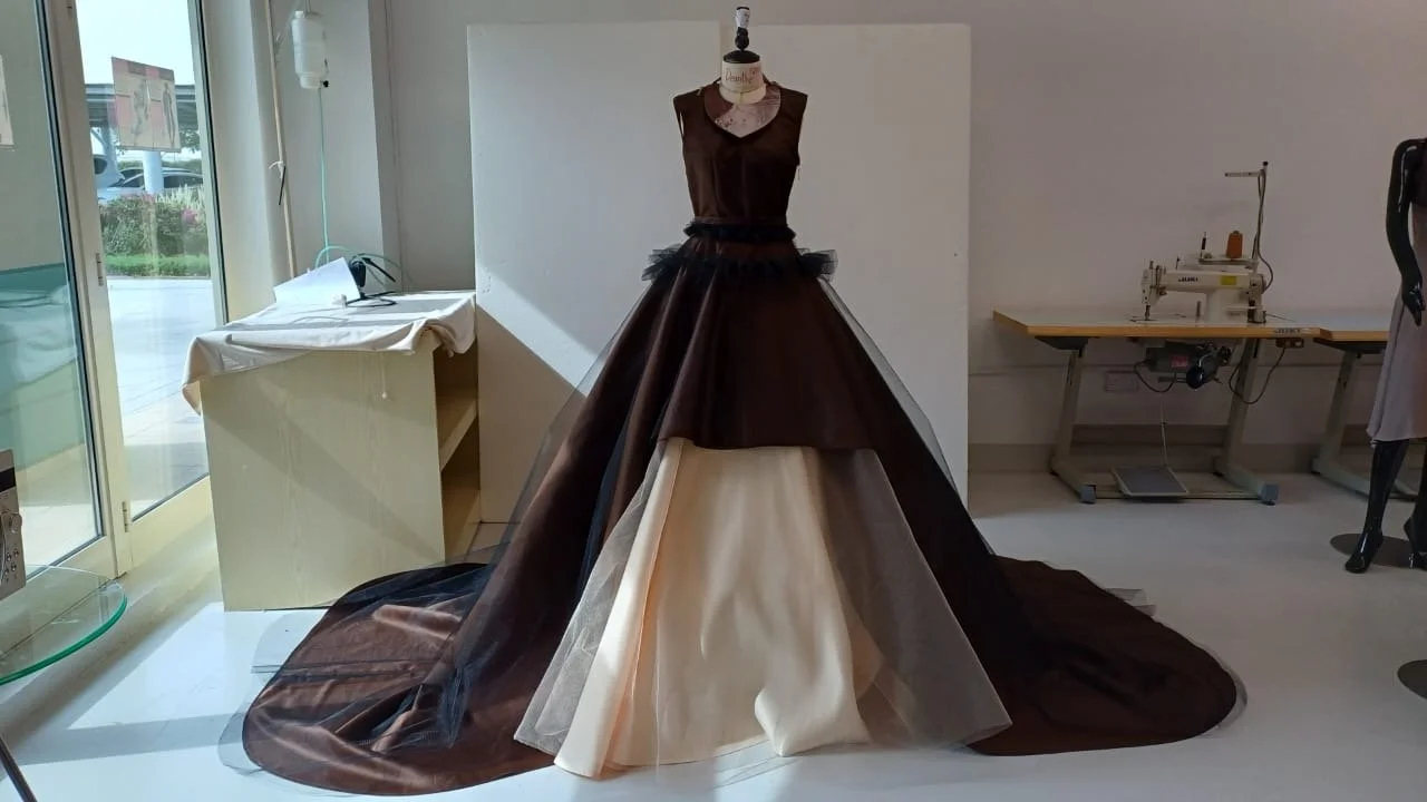 A mannequin dressed in a formal gown with a dark brown top, black tulle waist, and a layered skirt with white, beige, and brown fabric