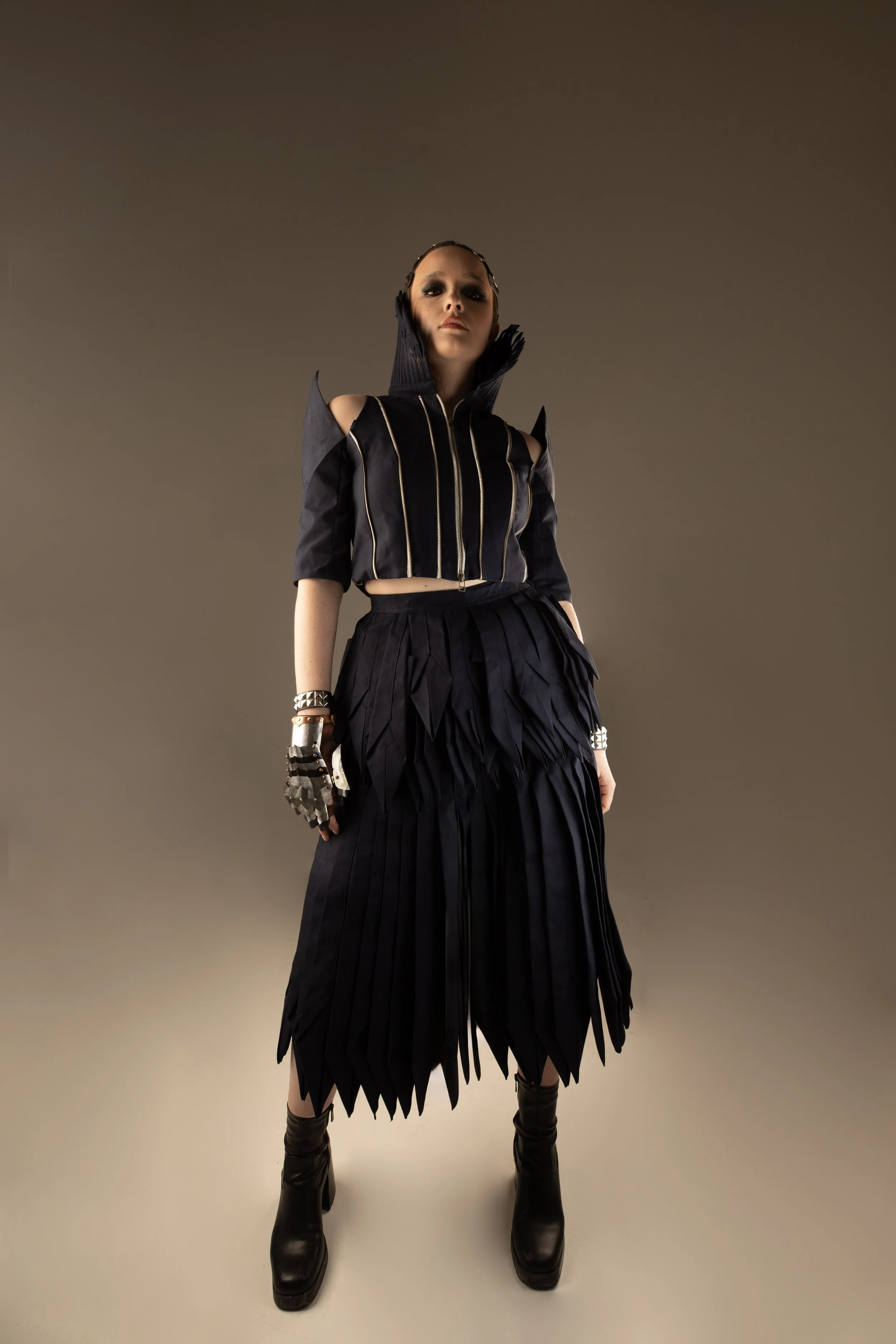 A woman wearing a black futuristic costume with pleated skirt, gloves, and high-heeled boots, standing against a neutral background.