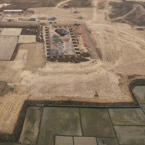 An aerial view of a rectangular construction site in a rural area, surrounded by open fields and dirt roads.
