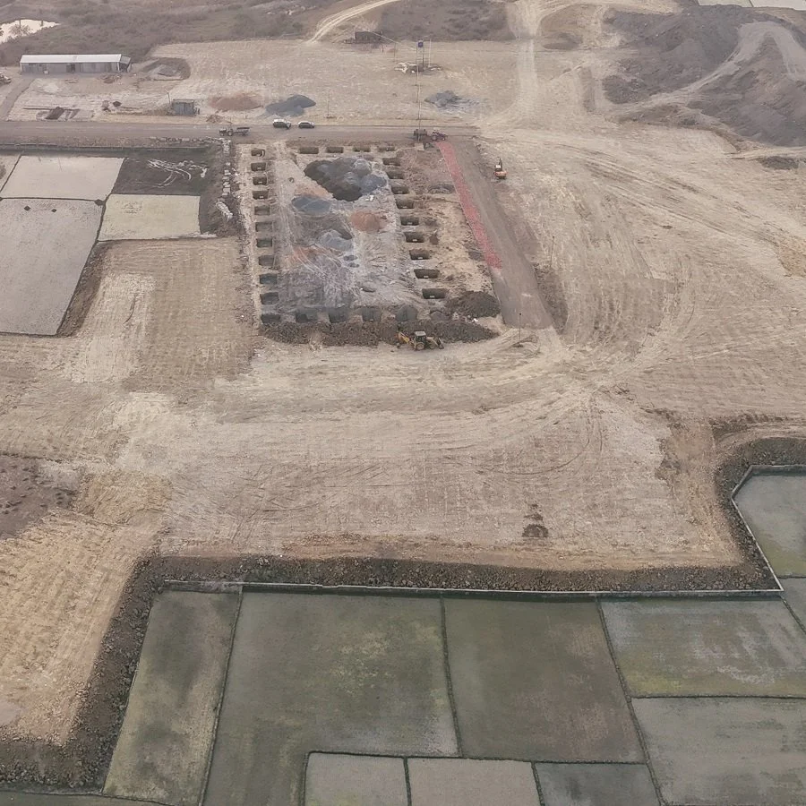 Construction site with trenches, dirt piles, and excavators, surrounded by fields and dirt roads.