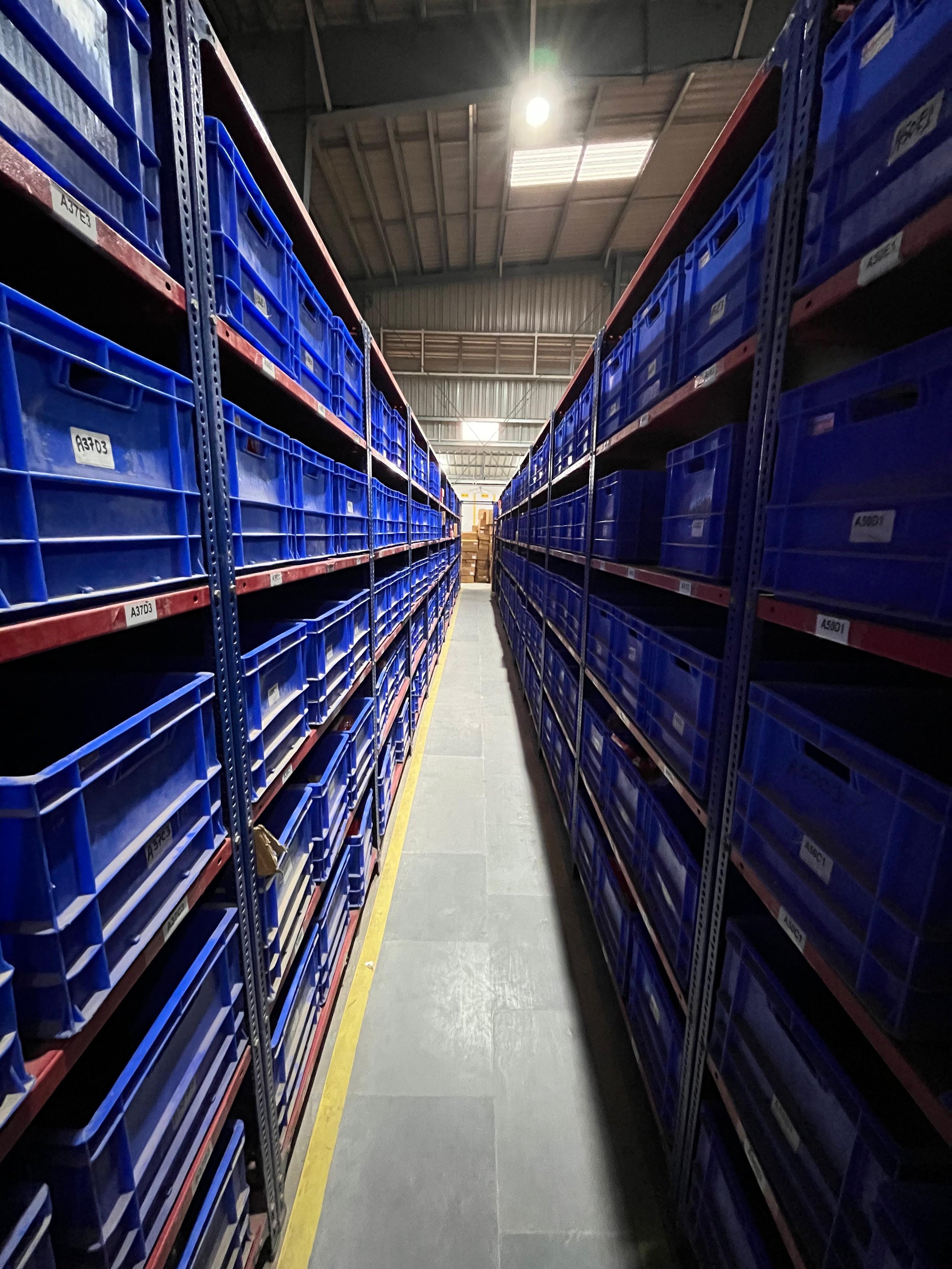 A narrow aisle in a warehouse with tall shelves stocked with blue storage bins on both sides.