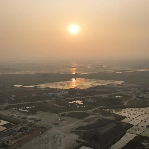 Aerial view of a landscape at sunset with water bodies, agricultural fields, and roads, under a hazy sky with the sun low on the horizon.