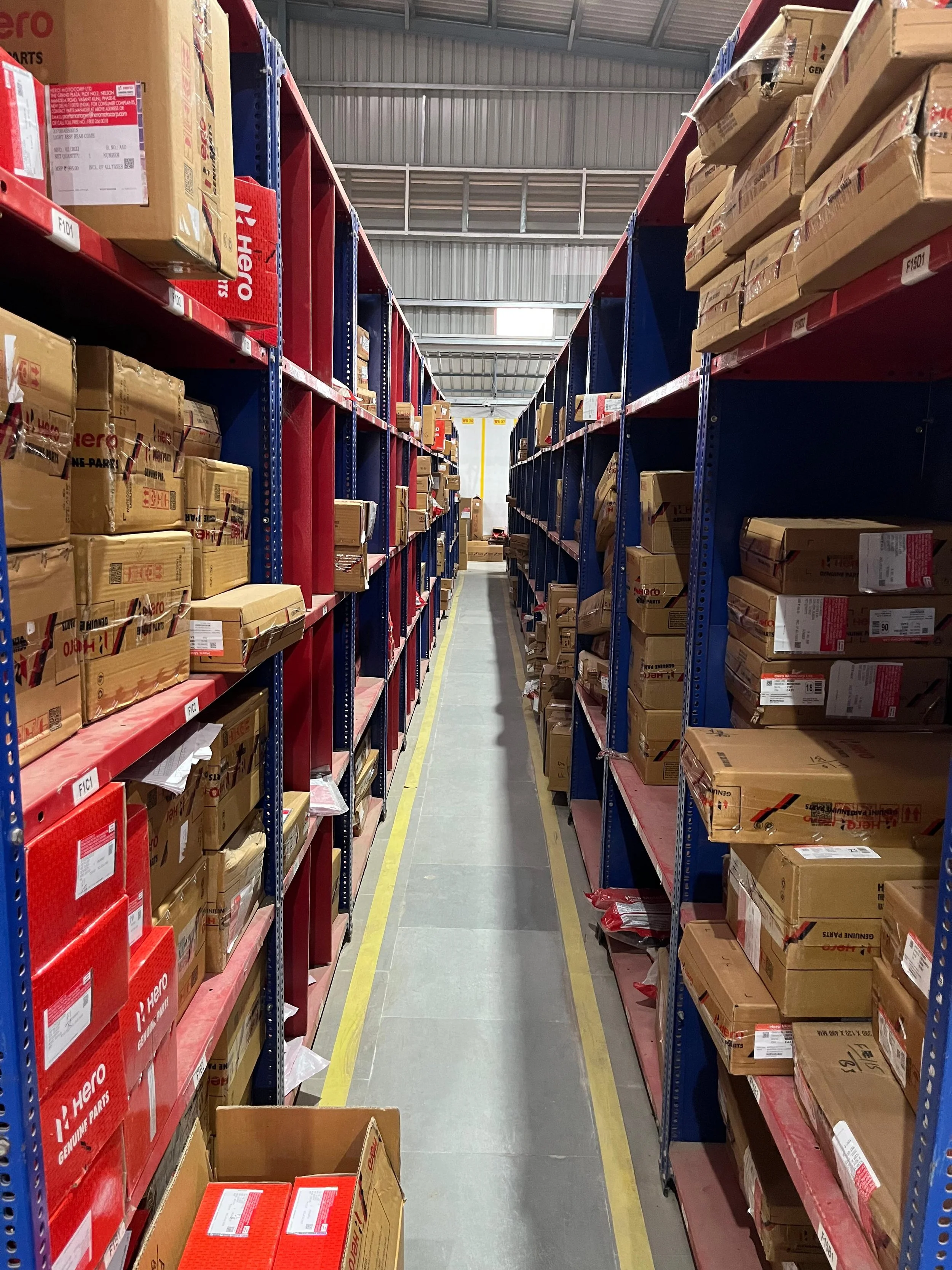 A warehouse aisle with shelves filled with brown boxes, some red boxes, and labeled with product information, extending into the distance.