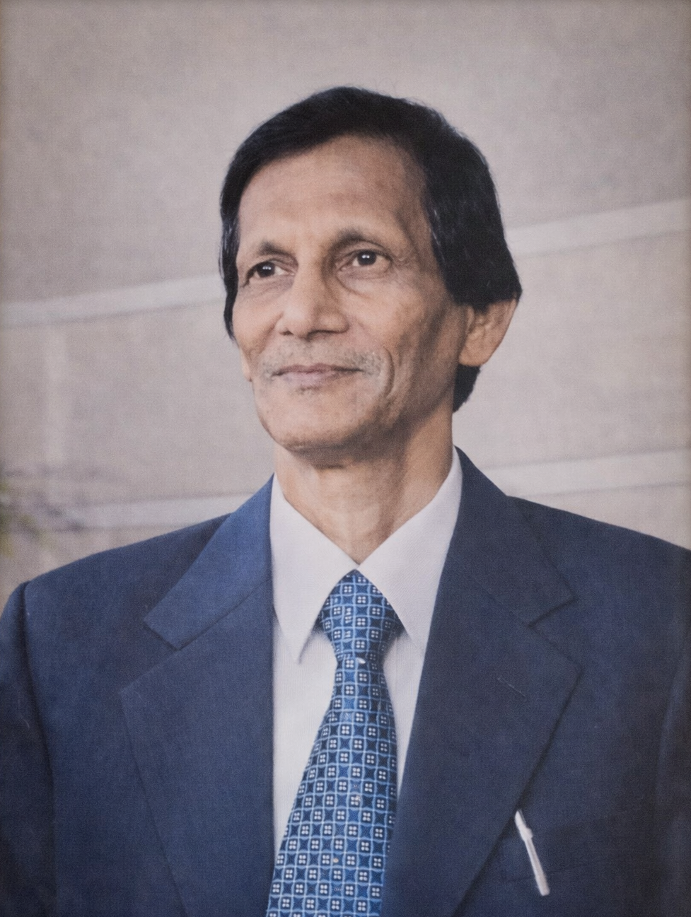 A man in a dark blue suit with a patterned tie, standing against a plain background.