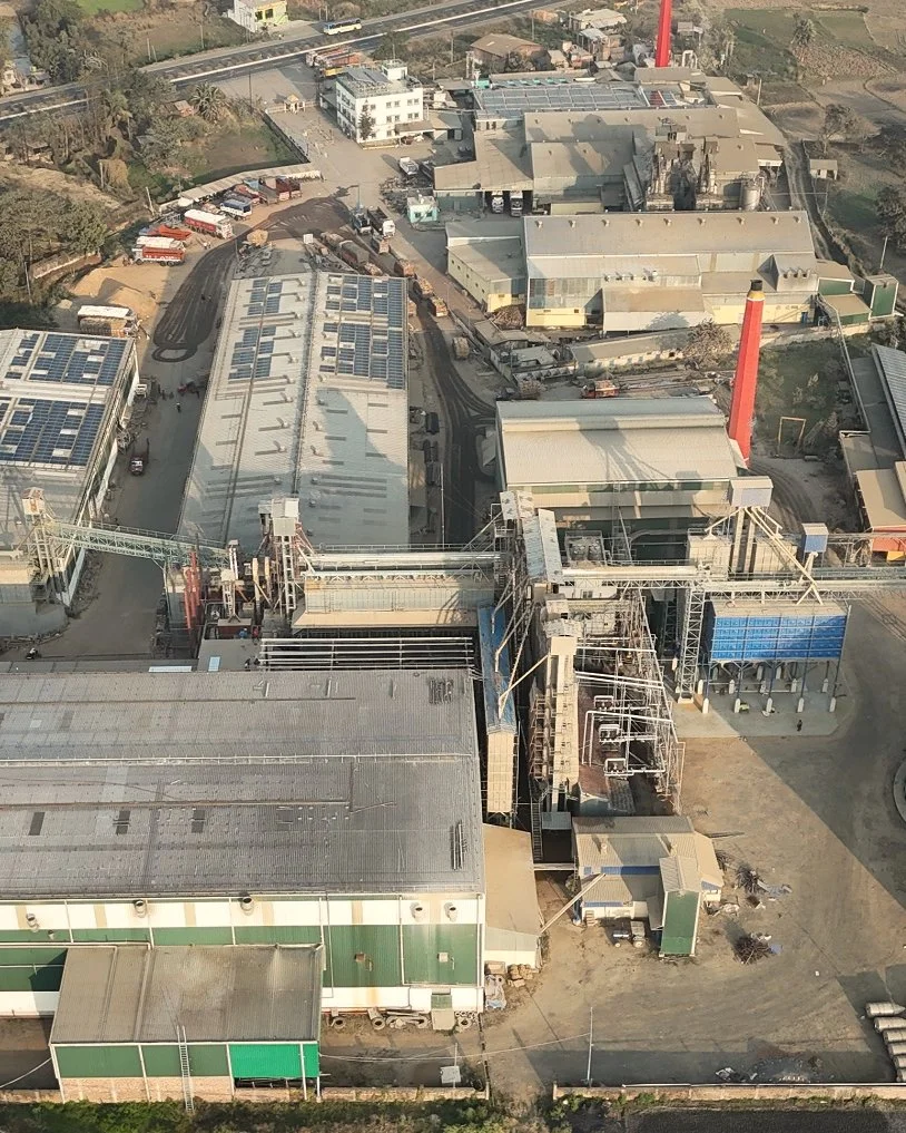 An aerial view of an industrial facility with multiple buildings, storage tanks, a red chimney, and solar panels on one roof. Tracks and vehicles are visible around the buildings.