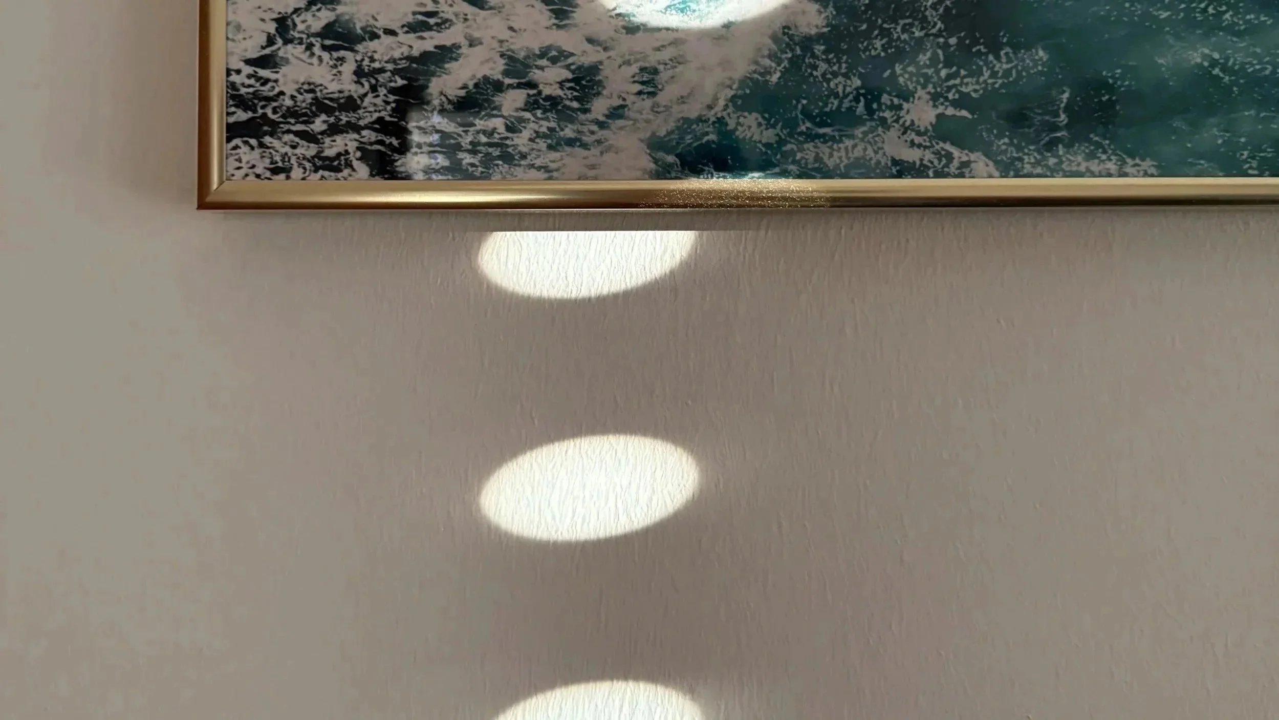 Part of a framed ocean or wave artwork hanging on a beige wall, with three oval spots of bright light reflection on the wall below the frame.