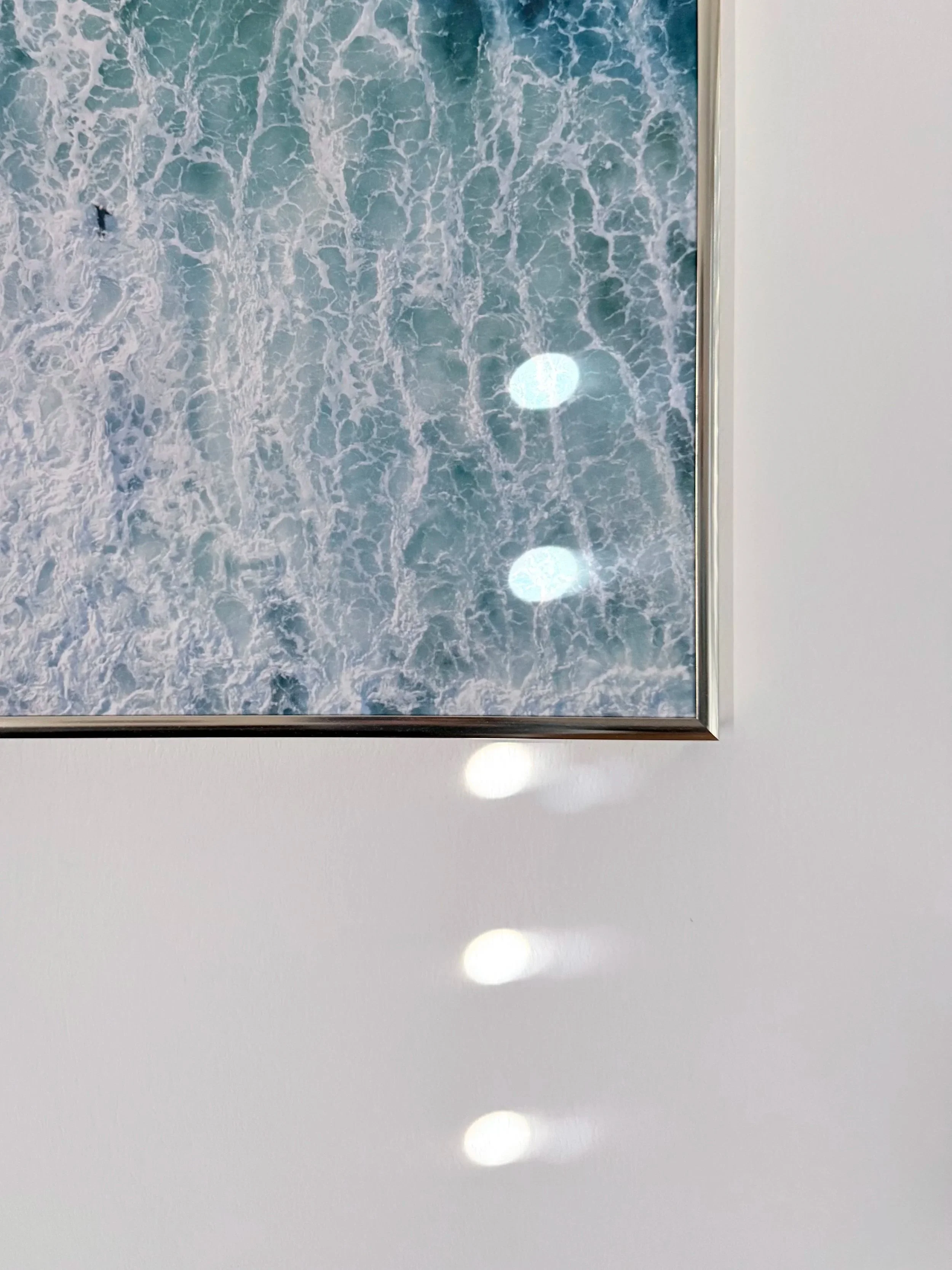 A framed photograph of ocean waves, showing white foam and blue water, hanging on a white wall with reflections of ceiling lights.