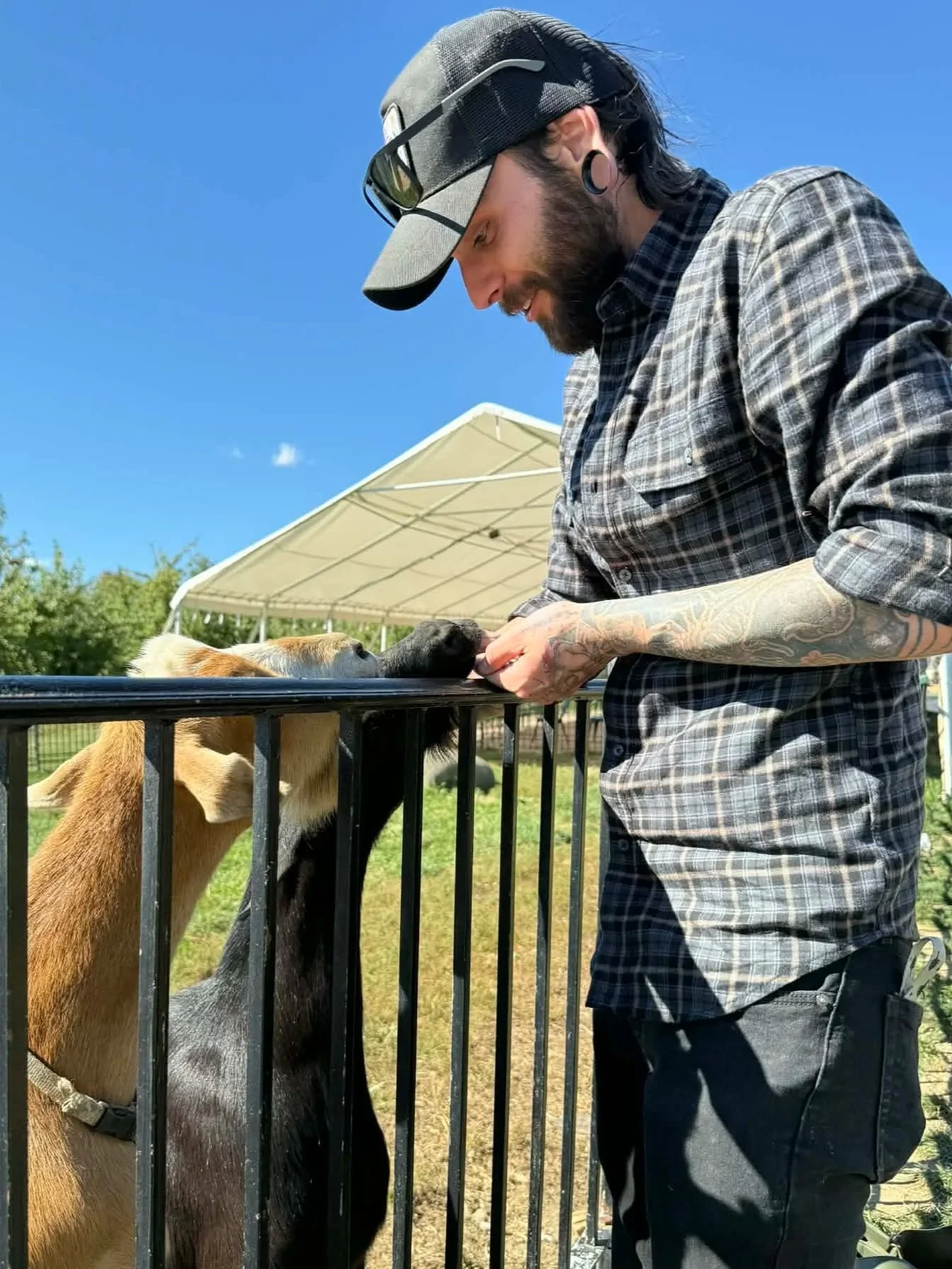 A man with glasses, a beard, and a nose ring is holding a large bird, possibly a wild turkey, close to his face. The man is wearing a black hoodie and a backpack. The background shows some trees and outdoor setting.