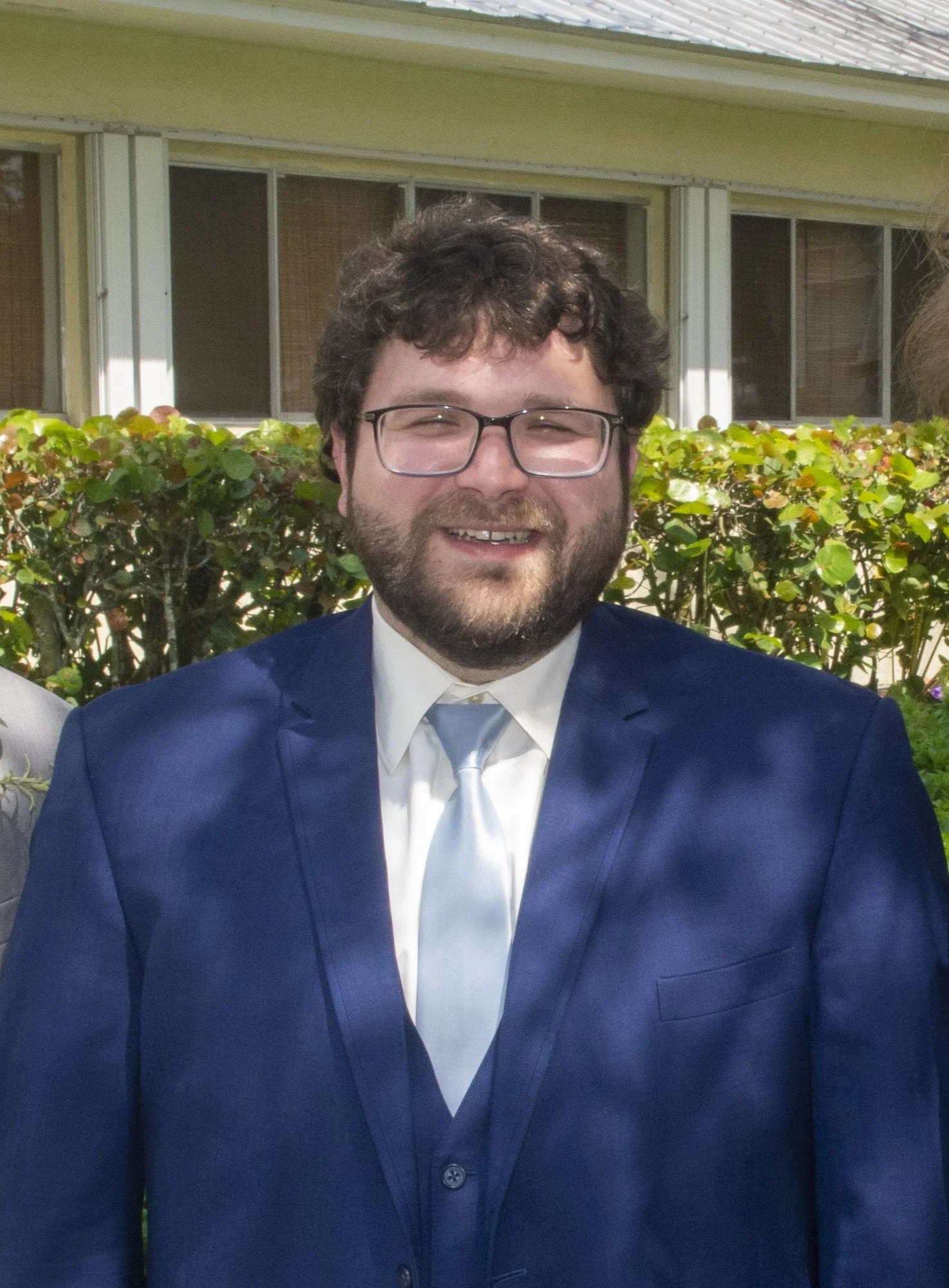 A man with curly hair, glasses, and a beard smiling, dressed in a navy blue suit, white shirt, and light blue tie, standing outdoors in front of bushes and a house.