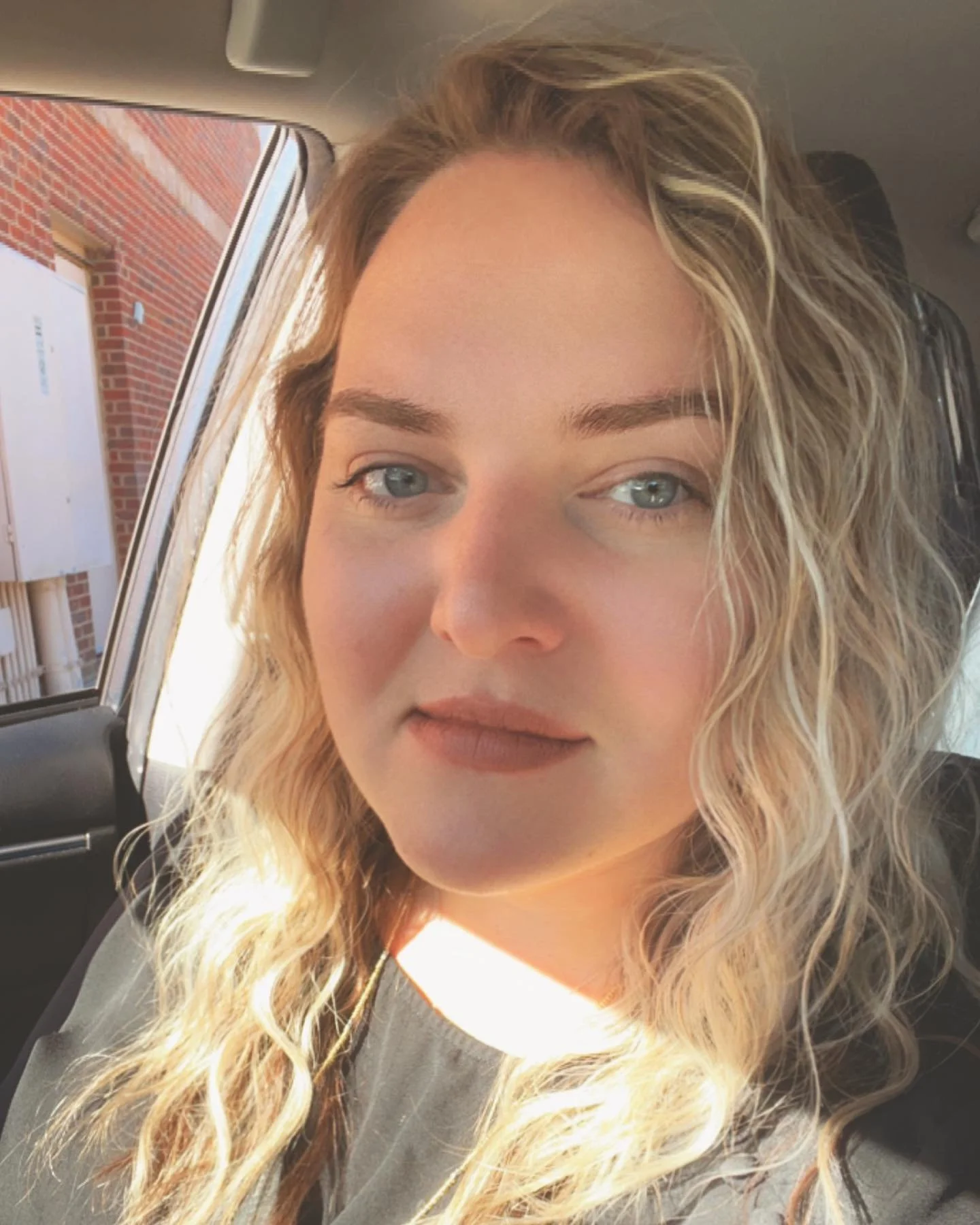 Close-up of a young woman with wavy blonde hair, wearing glasses, inside a car, with earbuds in her ears.