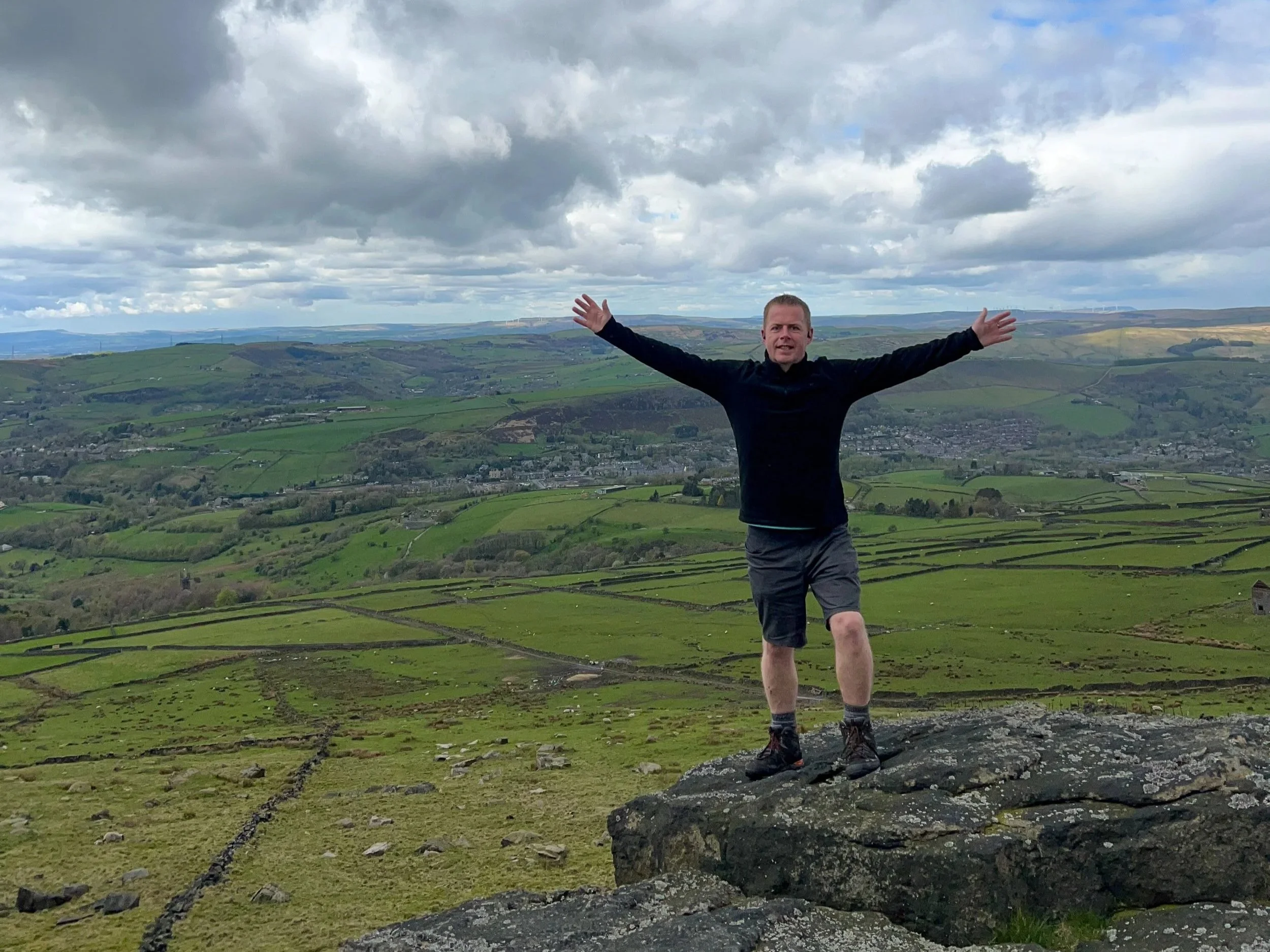 A man standing with arms outstretched on a large rock in a green, hilly landscape under a cloudy sky.
