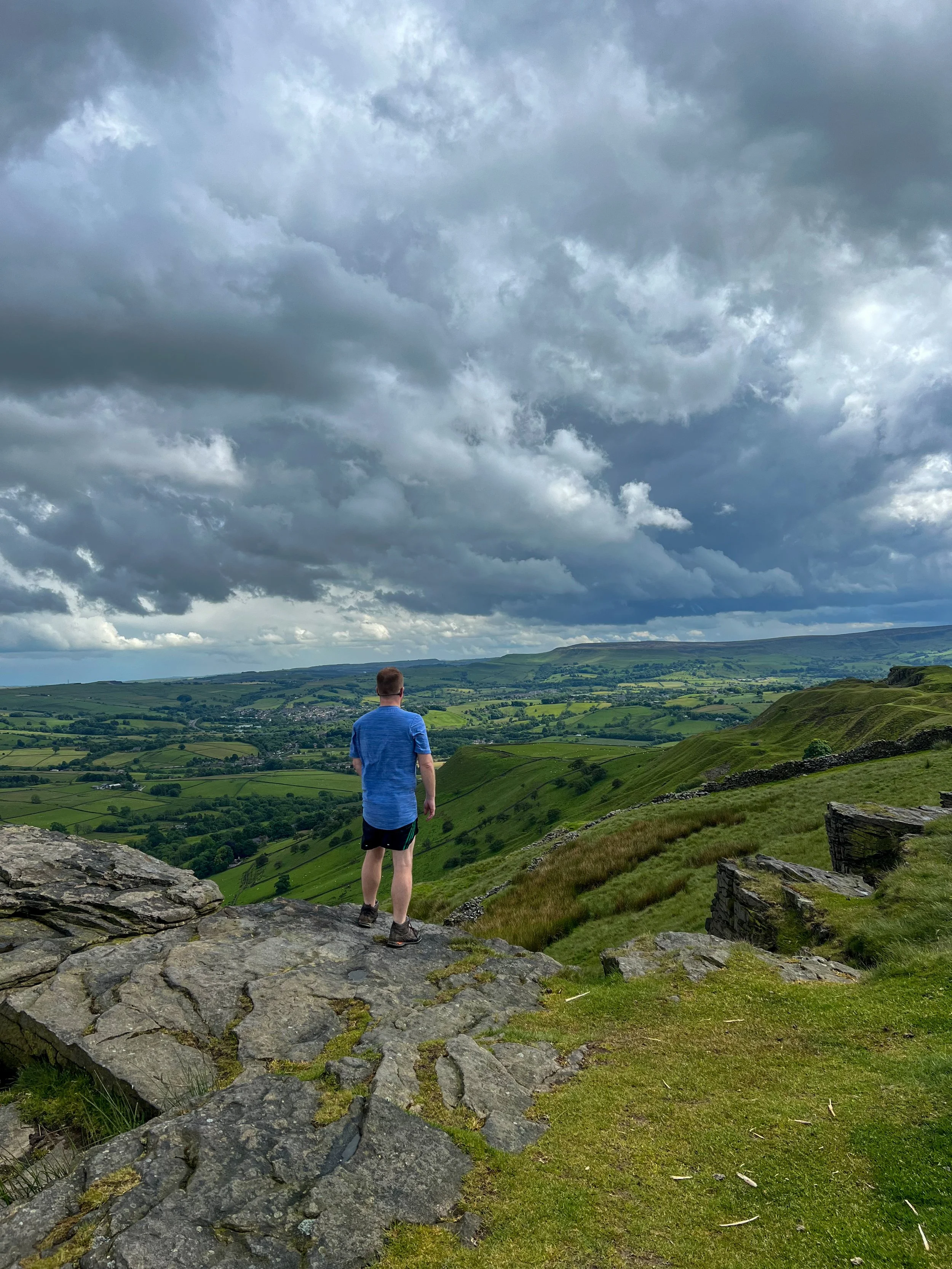 A man in a blue shirt and black shorts standing on a rocky ledge overlooking rolling green hills and a cloudy sky.