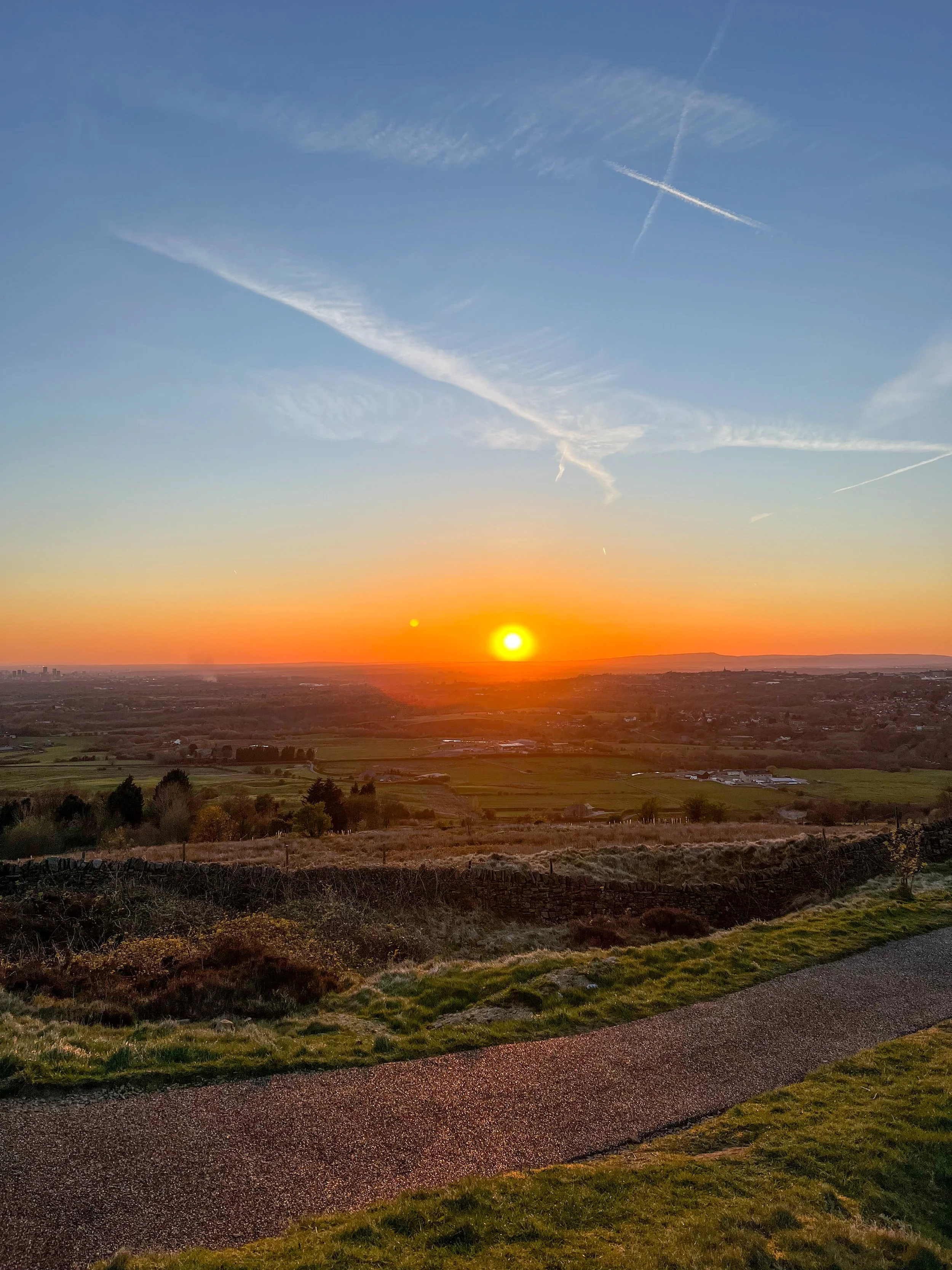 Sunset over a landscape with green fields, a stone wall, and a partly cloudy sky with jet contrails.