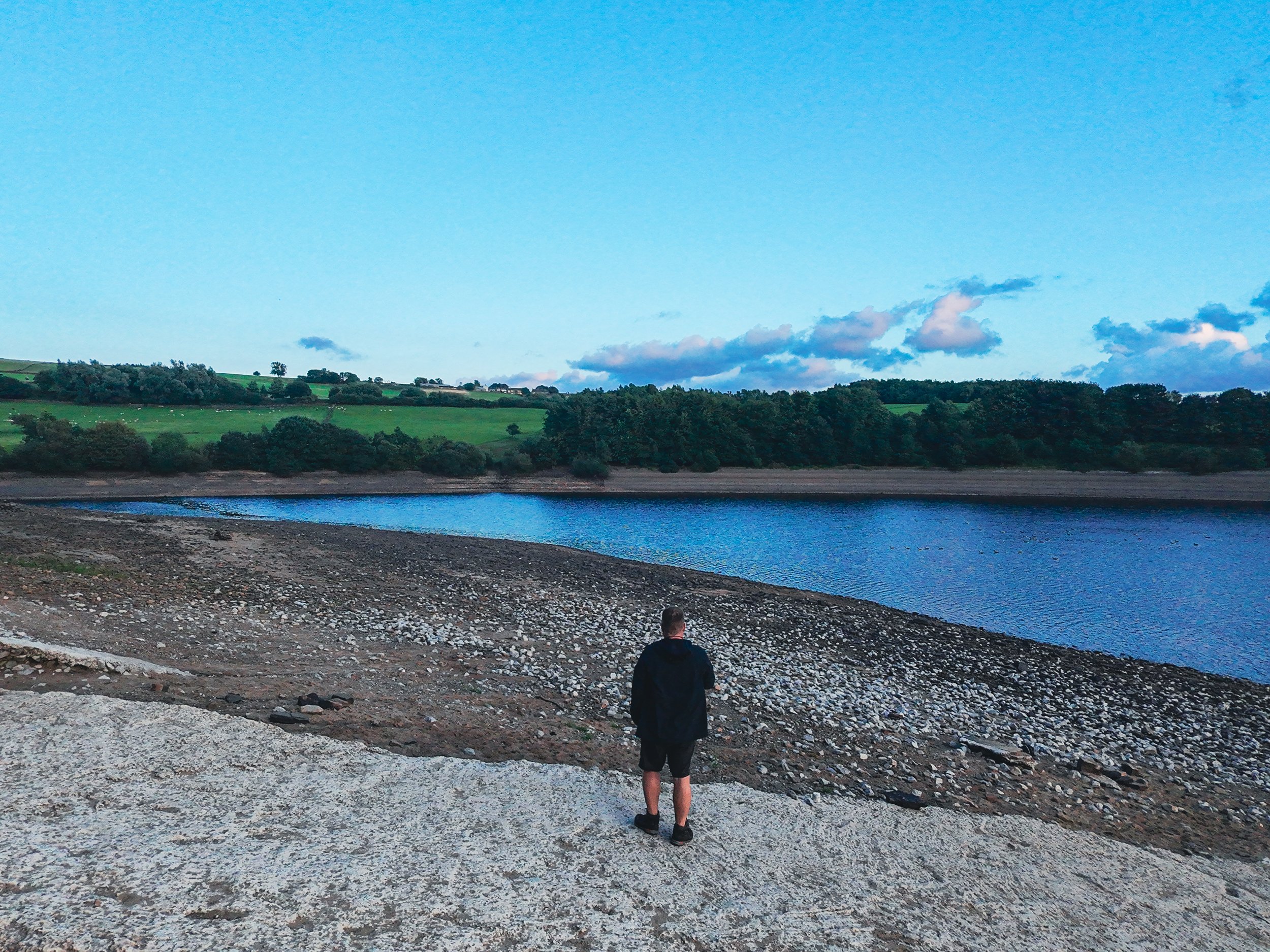 A person standing on a rocky, barren shoreline looking out over a body of water with green rolling hills and trees in the distance under a blue sky with some clouds.
