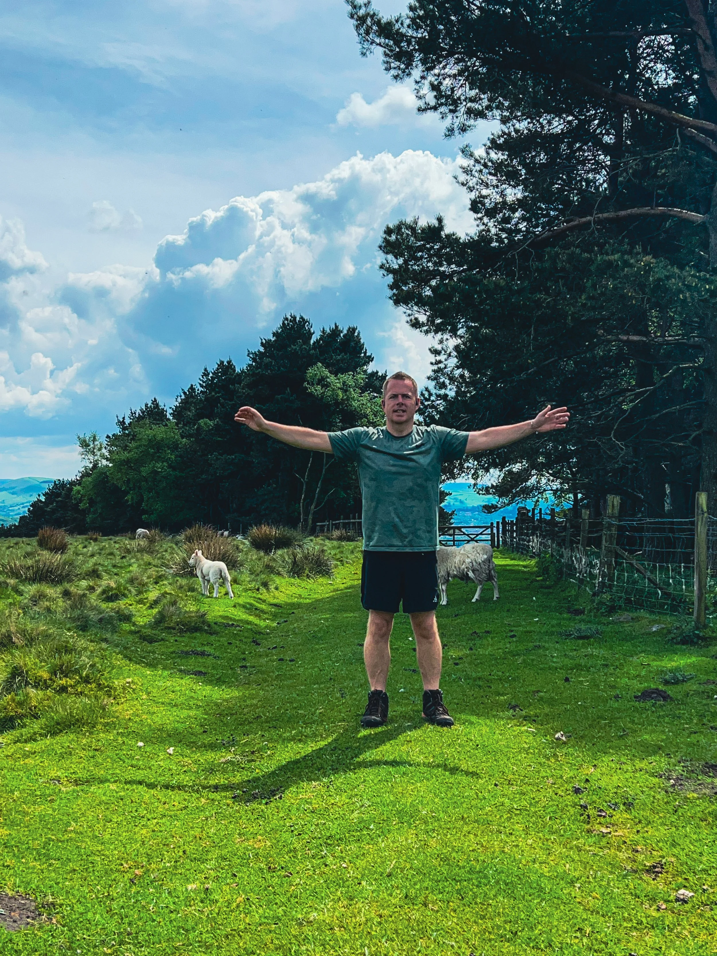 A man in athletic clothing standing with arms outstretched on a lush green path, with sheep and trees in the background under a partly cloudy sky.