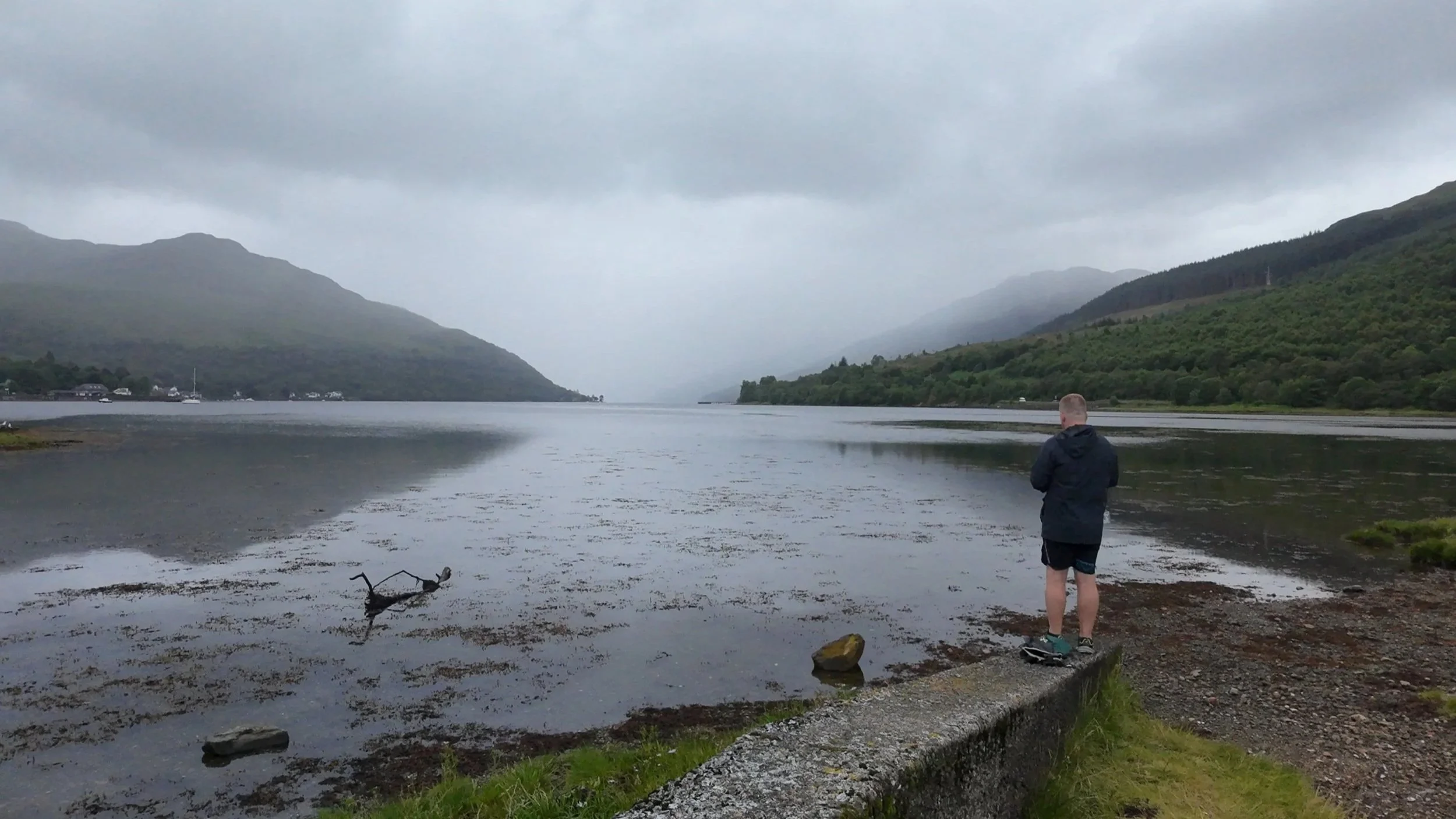 A young man in a dark jacket and shorts stands on the edge of a stone embankment, looking out over a calm, wide river surrounded by green hills and mountains under a cloudy sky.