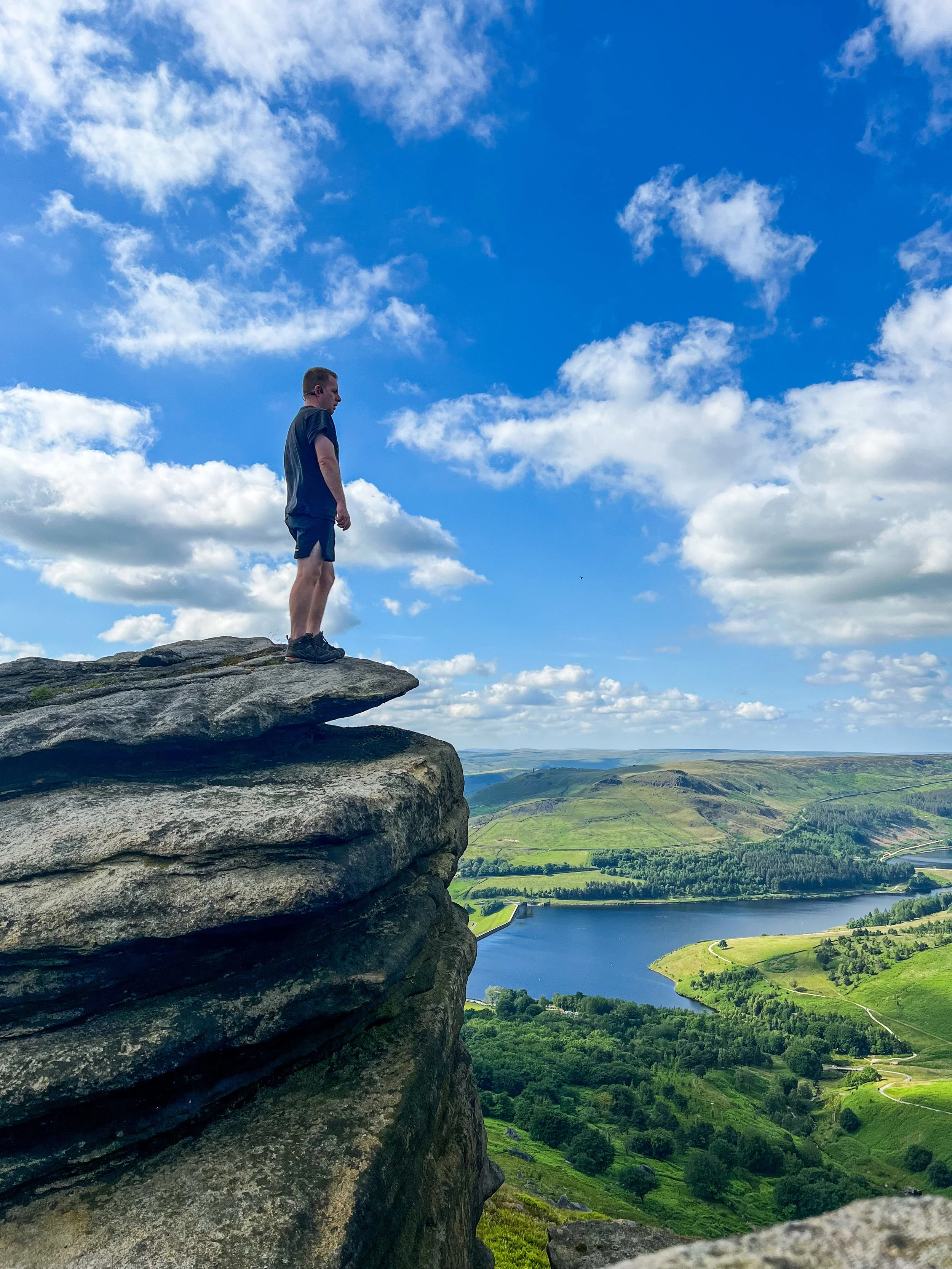 A man standing on the edge of a large rock formation overlooking a green landscape with a lake and cloudy sky.