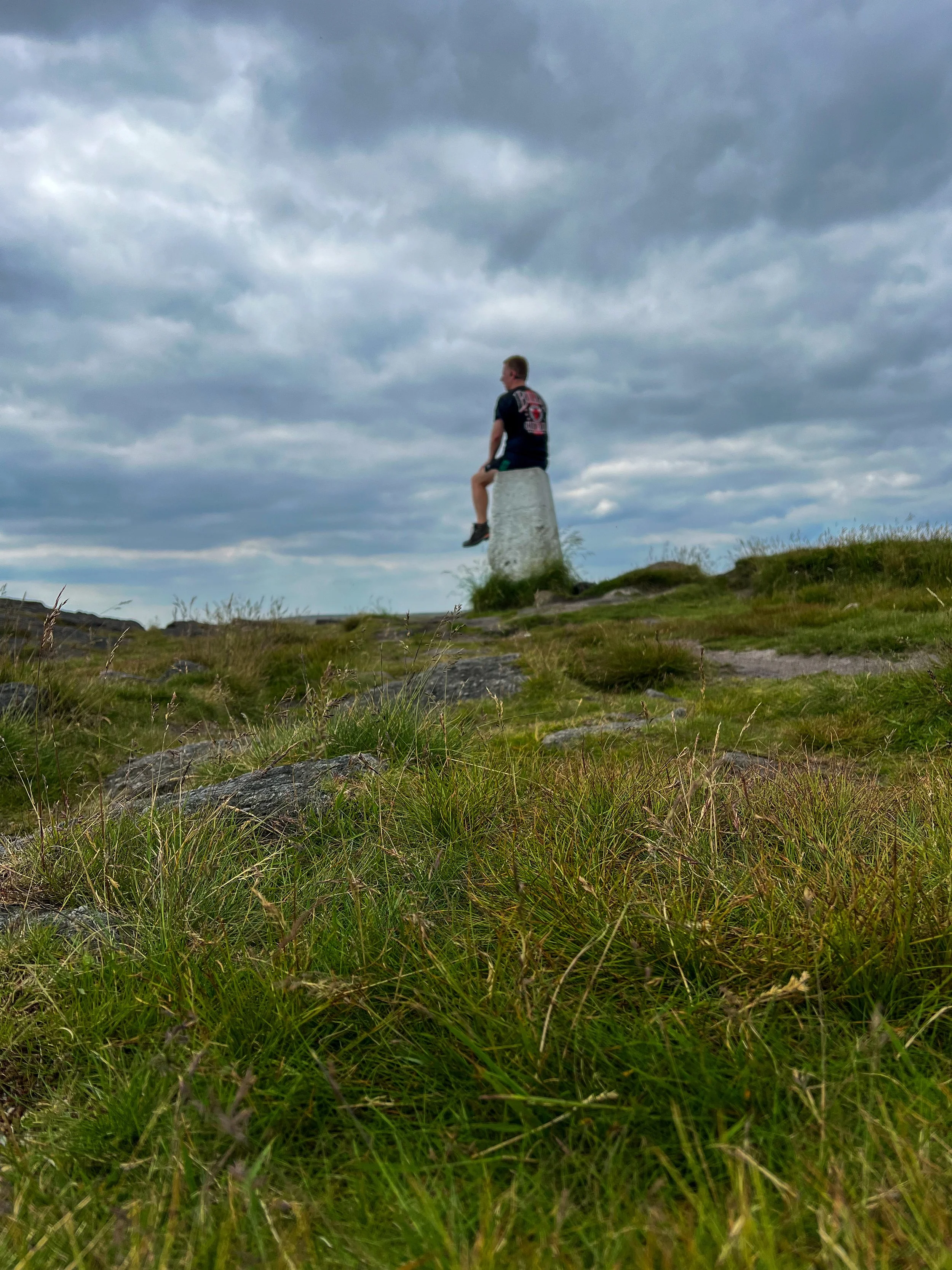 A person sitting on a concrete pillar outdoors against a cloudy sky, with grassy terrain and rocks in the foreground.