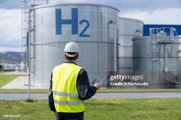 A person in a yellow safety vest and white helmet standing outdoors near large industrial tanks labeled 'H2' for hydrogen. The person is holding a clipboard and appears to be inspecting the facility.
