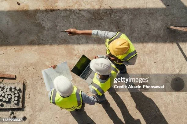 Three construction workers in safety gear discussing plans while looking at a laptop and blueprints on a construction site.