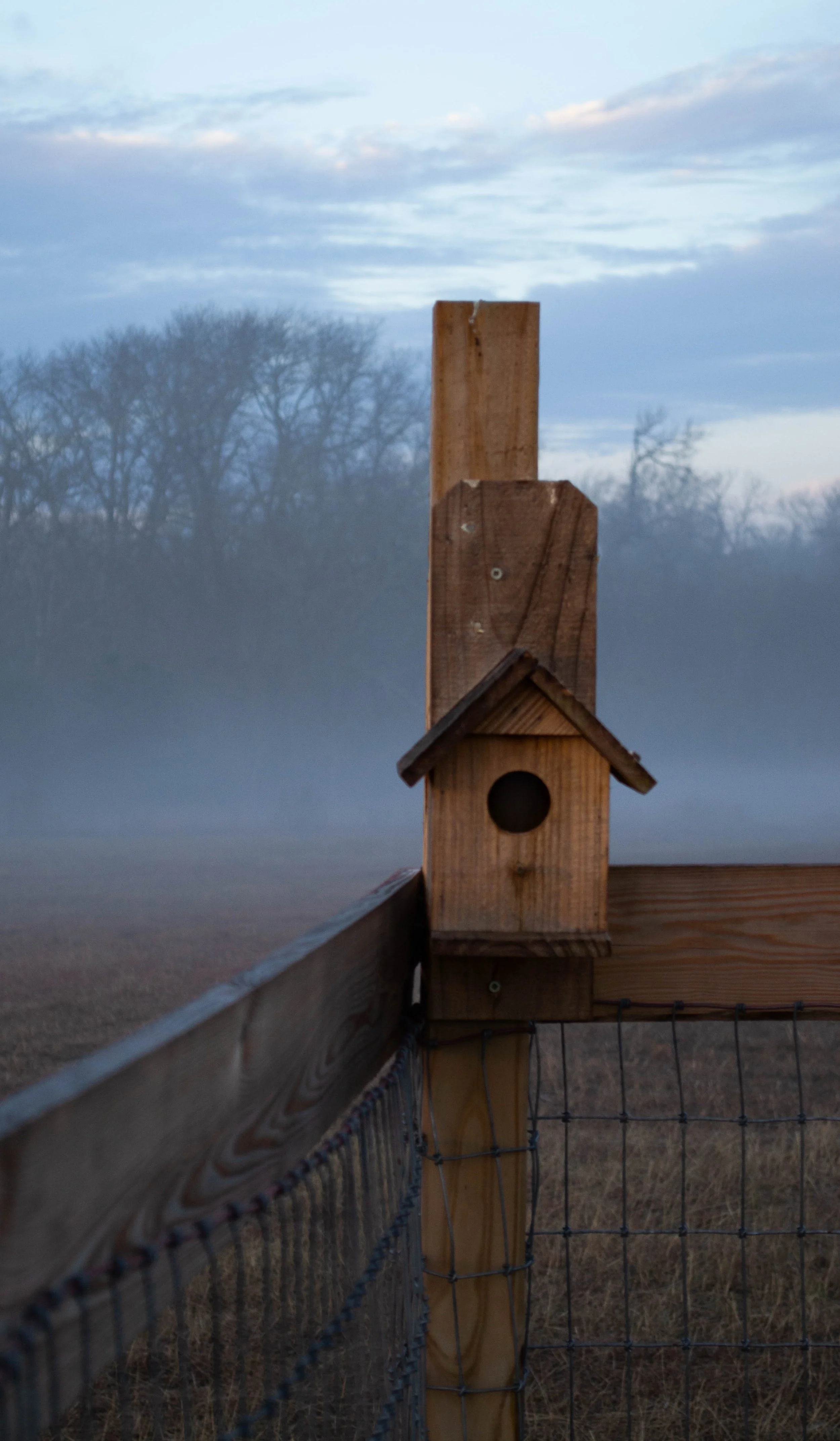 A small wooden birdhouse mounted on a wooden fence post, with a background of trees and a cloudy sky.
