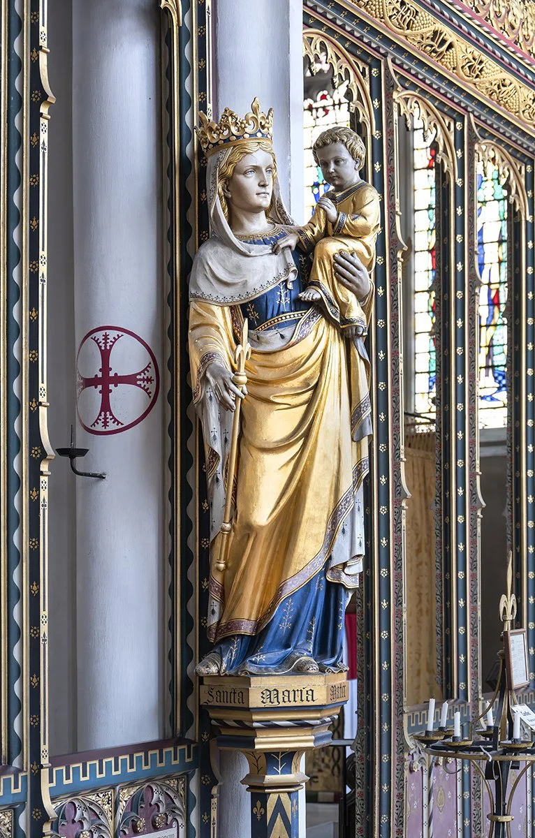 Religious statue of the Virgin Mary holding baby Jesus inside a church with stained glass windows in the background.