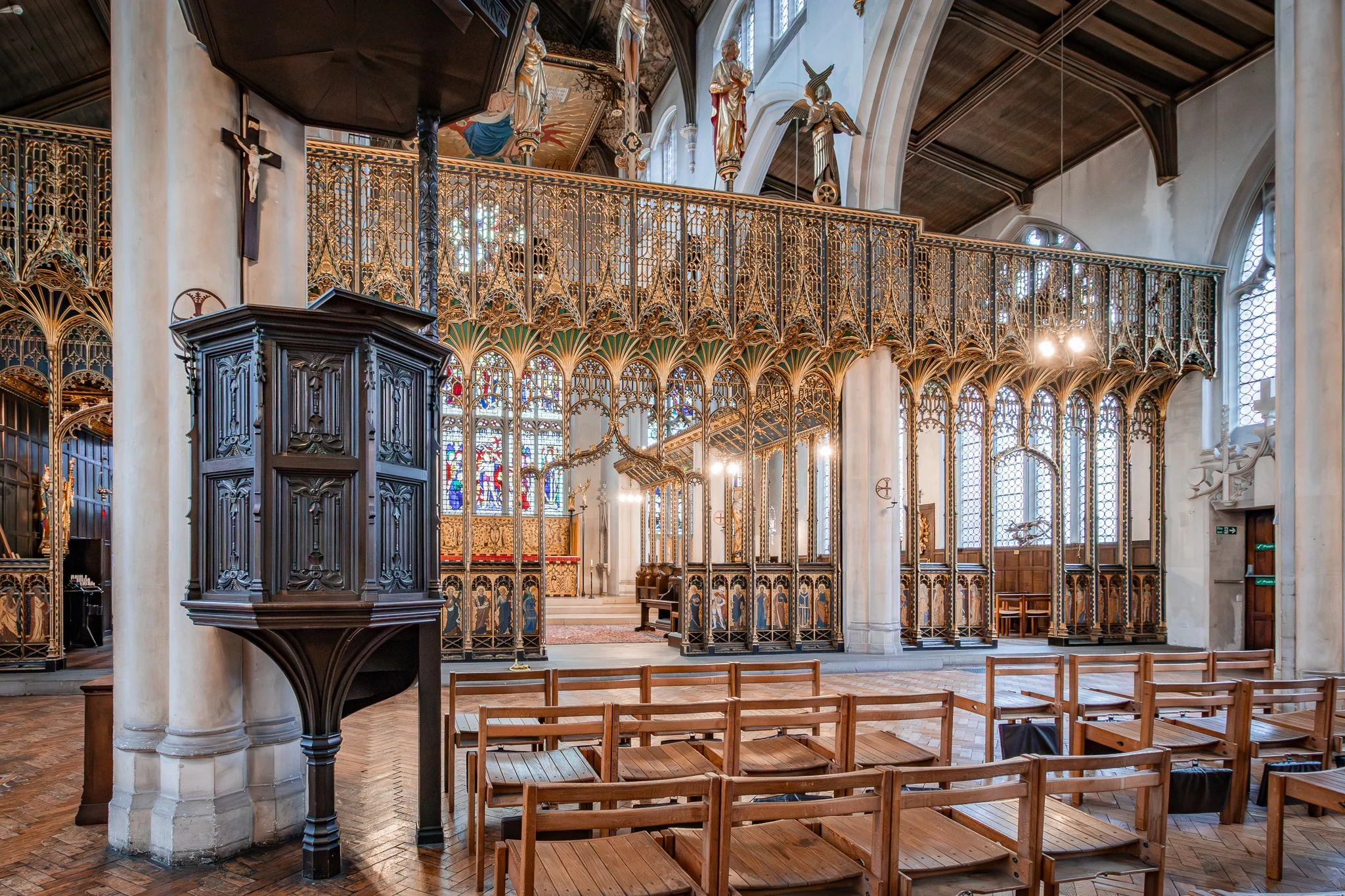 Interior of a church with wooden benches, stained glass windows, and ornate gold-colored ironwork separating the chancel from the nave.