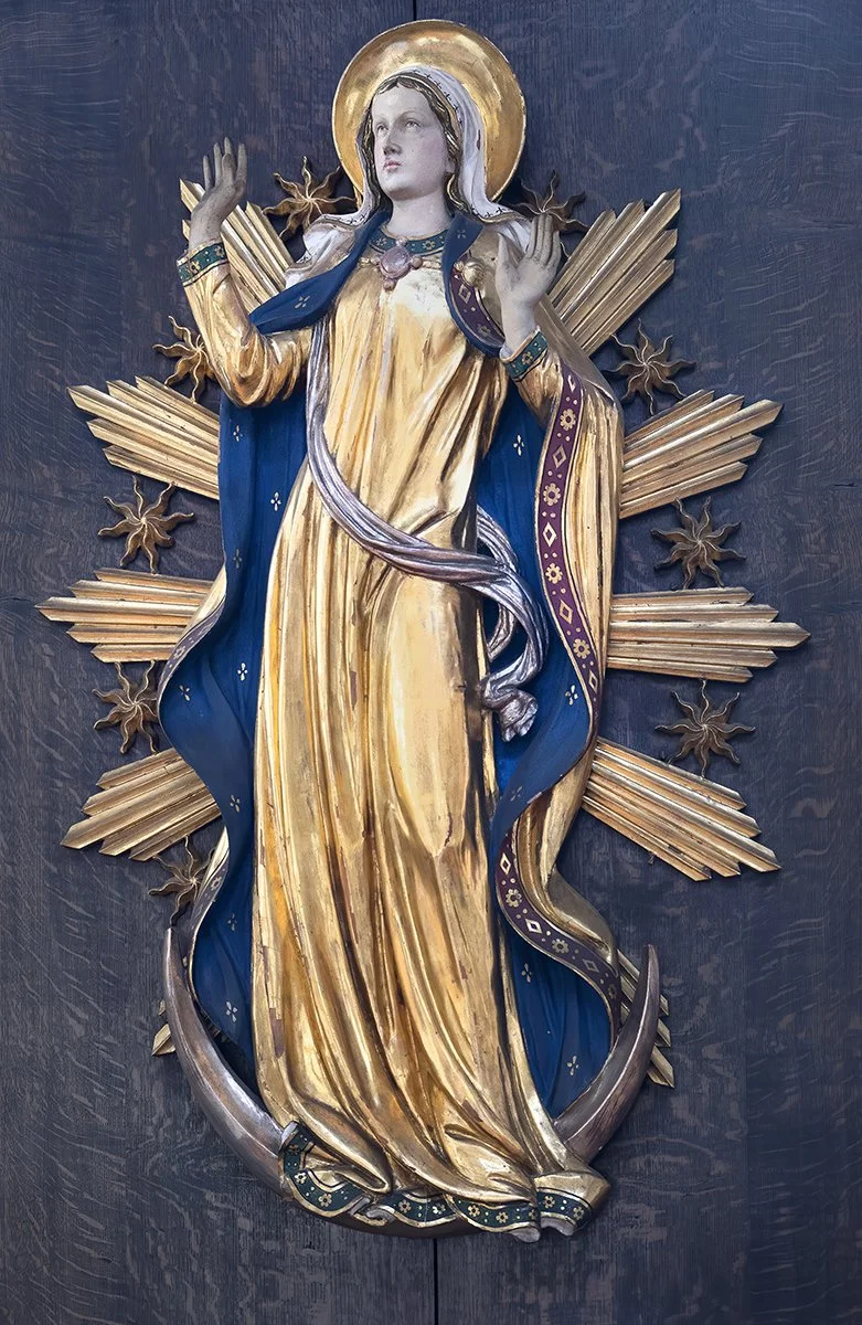 Religious artwork of a female saint or the Virgin Mary with a gold halo, dressed in a gold and blue robe, hands raised, standing against a dark background with gold rays and floral elements.