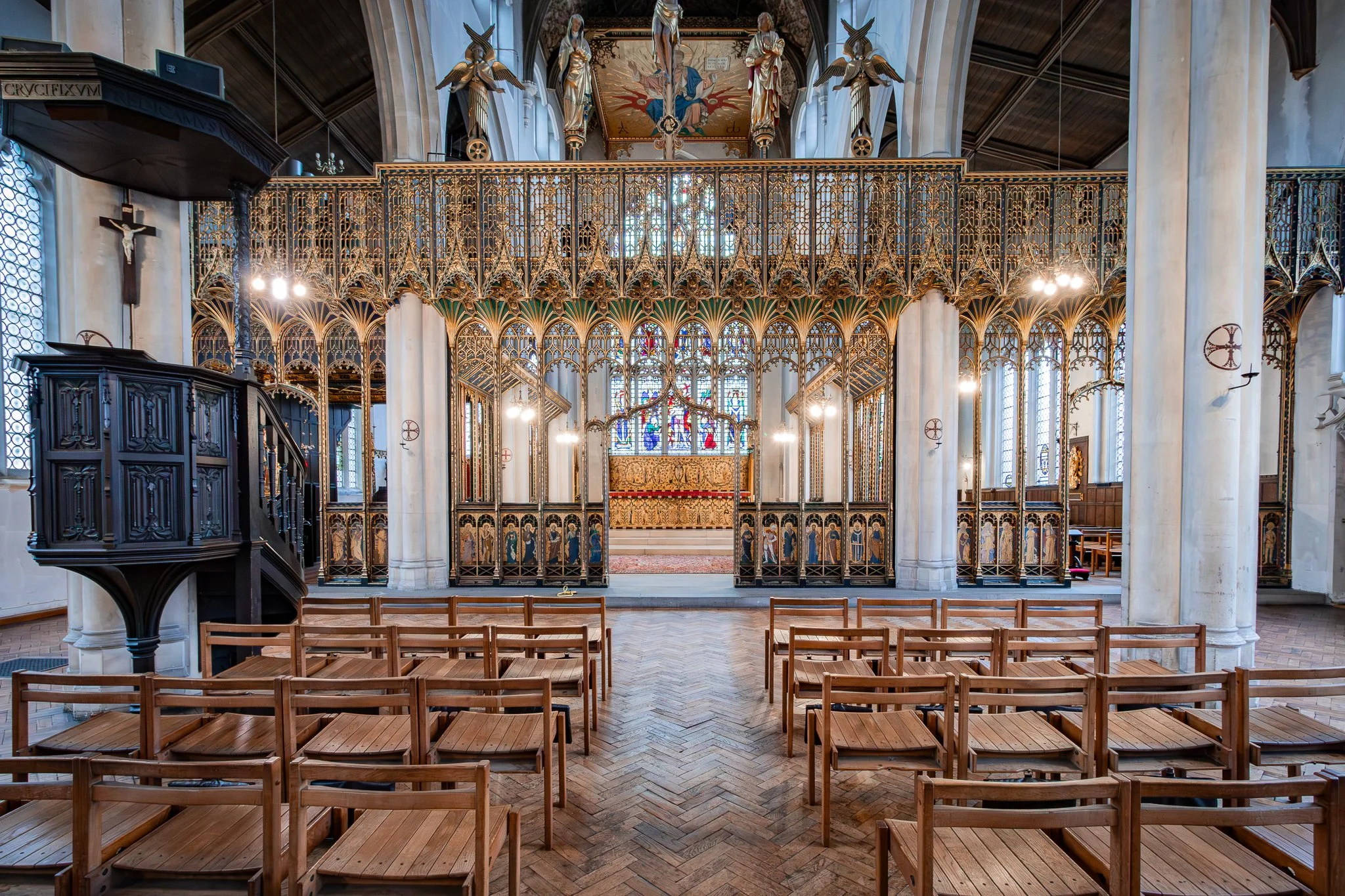 Interior of a church with wooden chairs arranged in rows, ornate gold detailing, stained glass windows, and religious symbols including a crucifix.