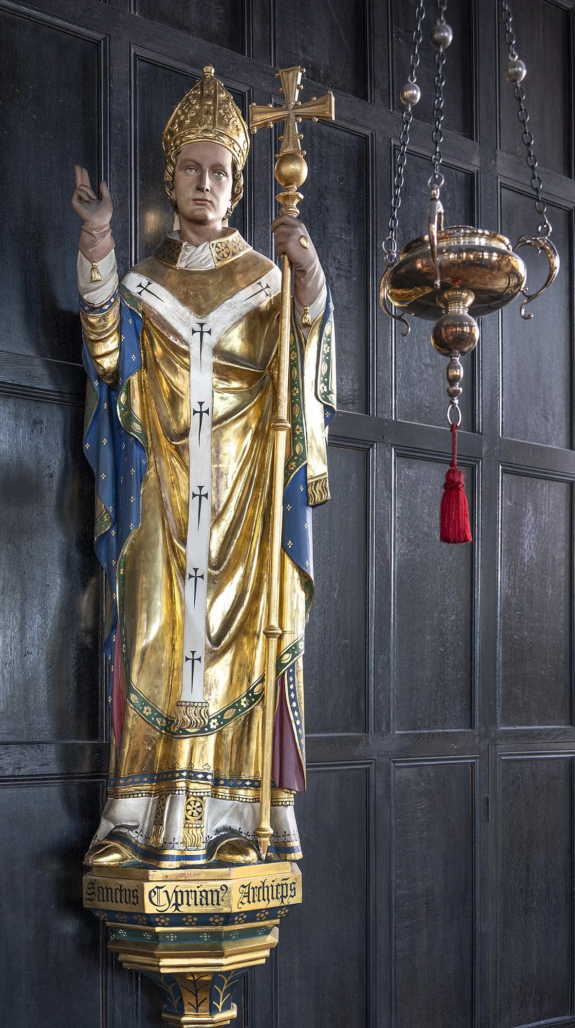A religious statue of Saint Cyrian Archangel dressed in gold and blue robes, holding a cross and a staff, mounted on a wall with dark wooden paneling.