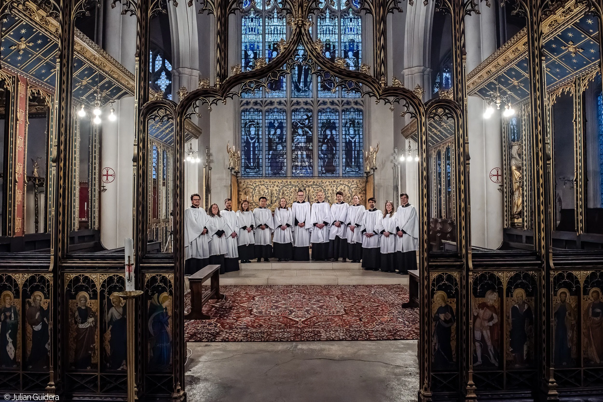 Choir of people dressed in white robes standing in a church or cathedral with stained glass windows and ornate decorations.