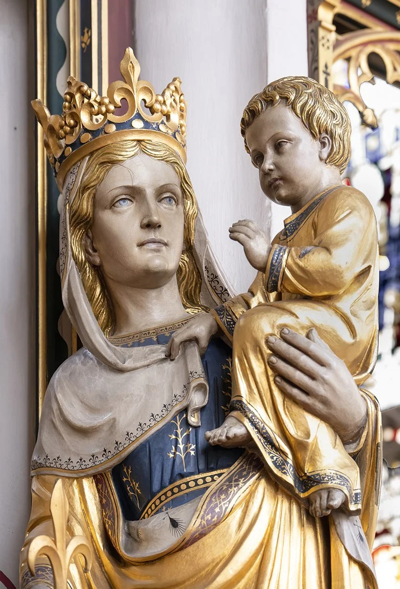 Religious sculpture of the Madonna and Child with gold accents, the Madonna wears a crown and a blue and white robe, holding the Child dressed in gold.