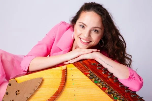 A smiling woman with curly hair, wearing a pink blazer, leaning on a harpsichord with a gray background.