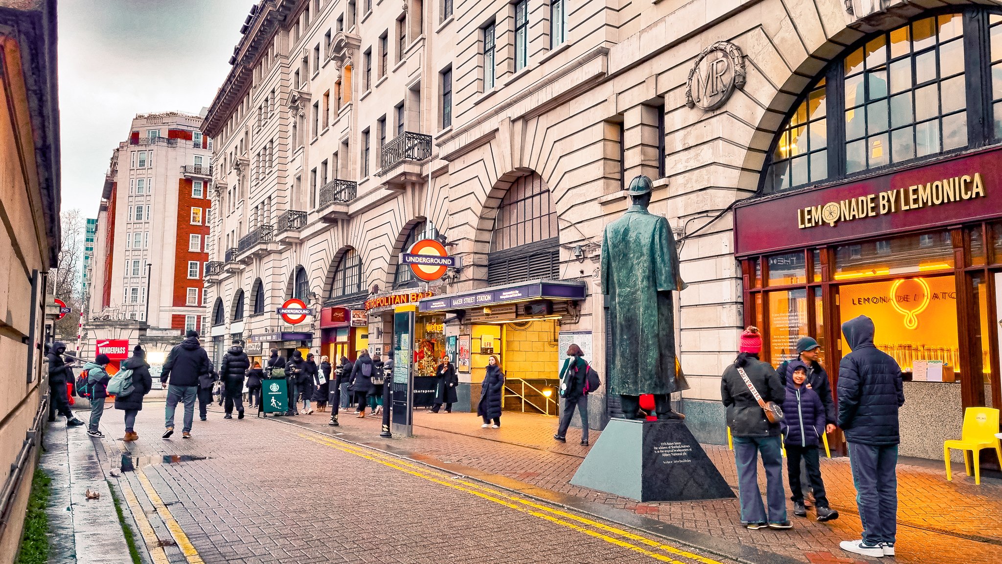 People waiting outside Baker Street Station, London, with a bronze statue of a man in a coat and hat in the foreground, and storefronts including 'Lemonade by Lemonica' on the right.