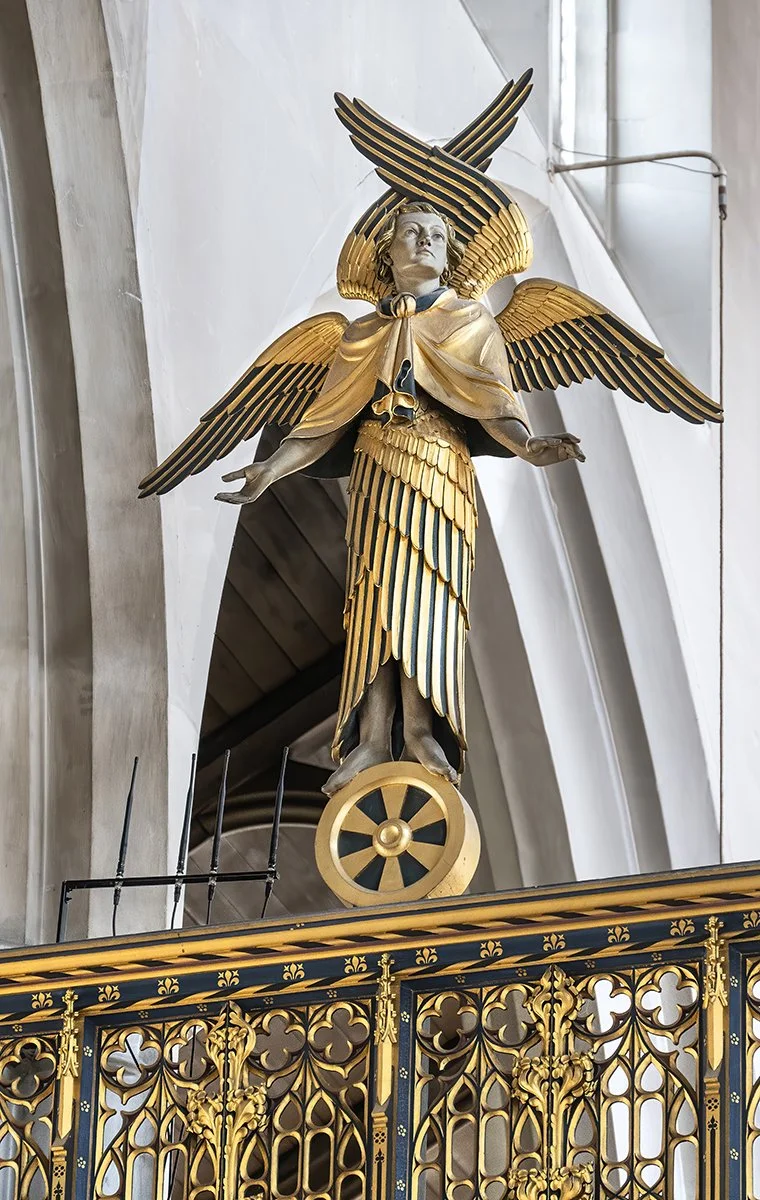 A golden angel statue with wings, wearing a golden robe and a cape, standing on a shield with a wheel at its base, inside a church.