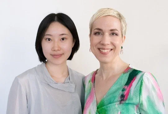 Two women standing side by side in front of a plain white background, smiling at the camera.