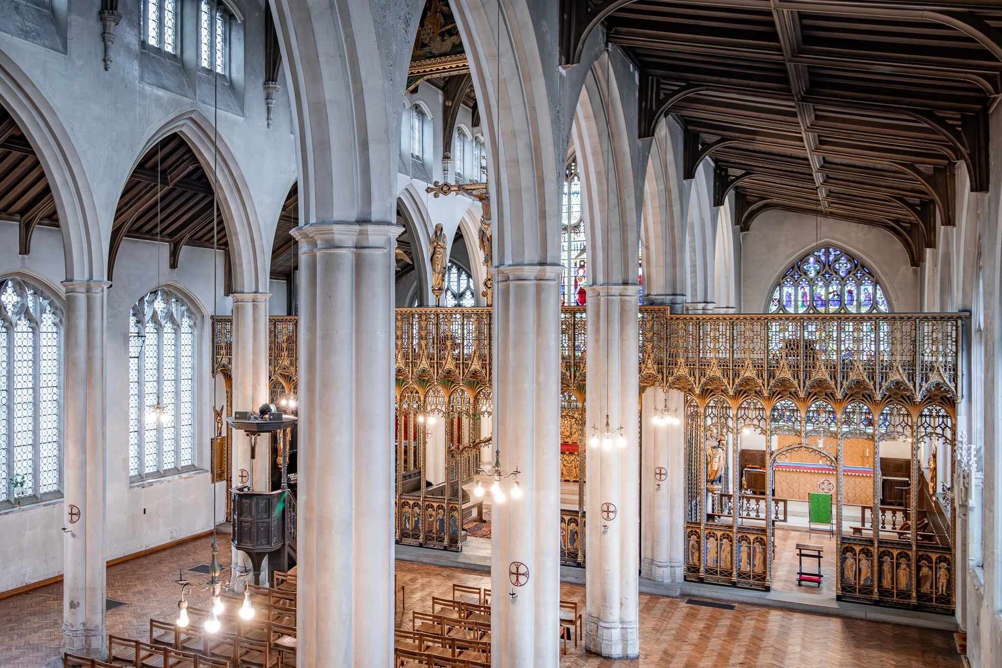 Interior of a Gothic-style church with tall stone columns, stained glass windows, wooden pews, and ornate gold railing.