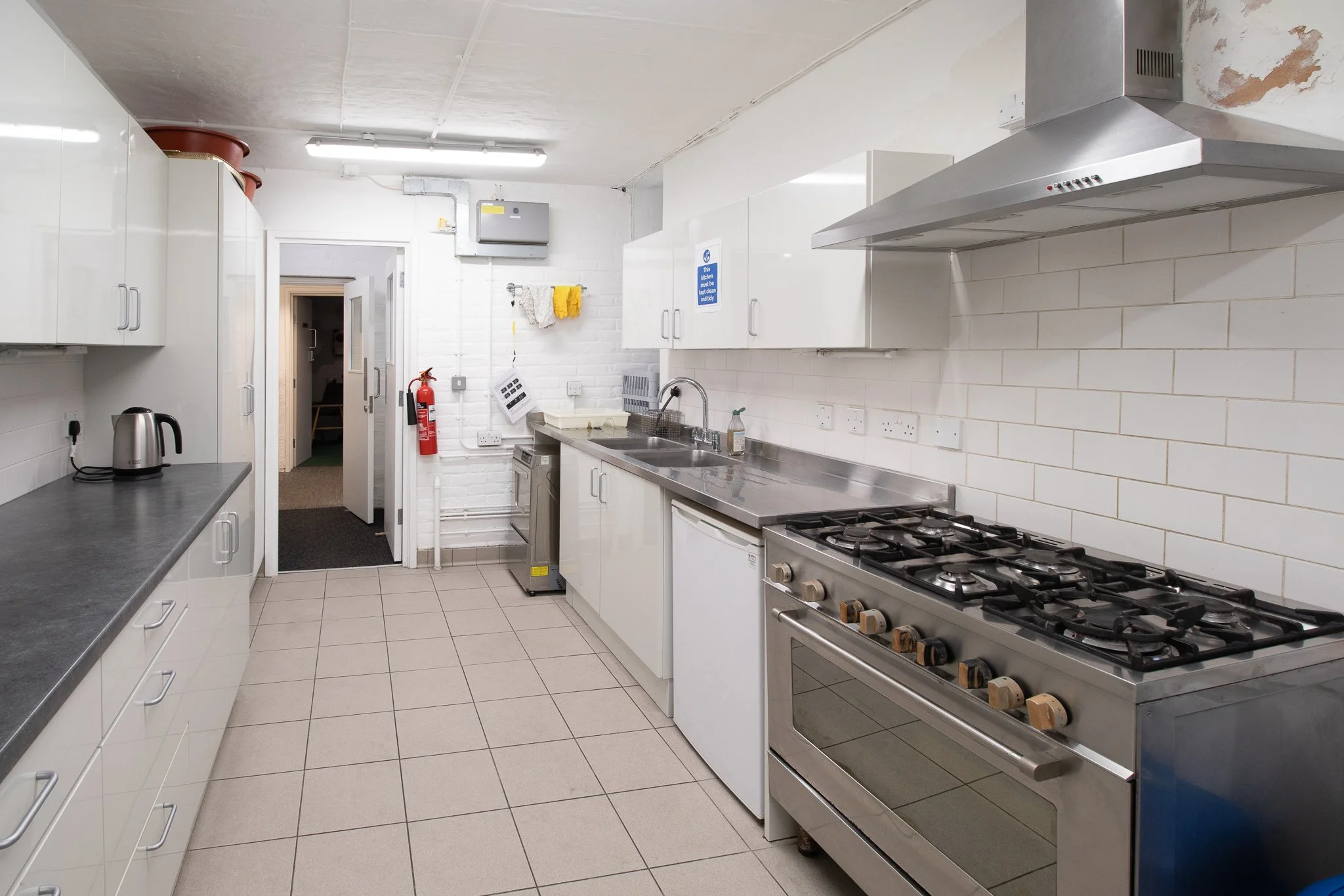 Kitchen with white cabinets, gray countertops, stainless steel appliances including a oven and range, a sink, white brick walls, tiled floor, and some items like pots, a kettle, and cleaning supplies.