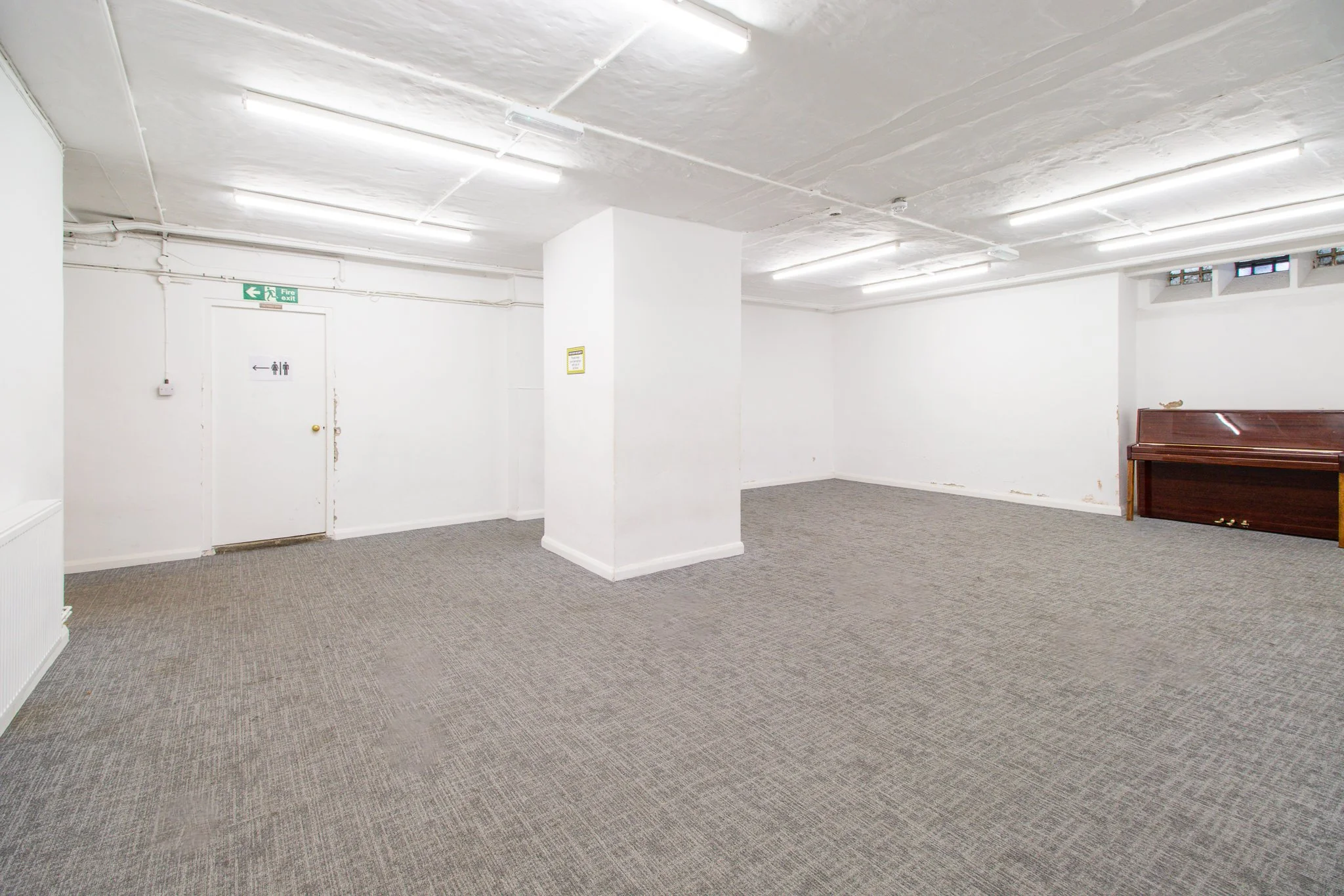 Empty room with white walls and gray carpet, small window near ceiling, wooden upright piano against right wall, ceiling with exposed pipes and fluorescent lights, fire exit door on left, small window near ceiling.