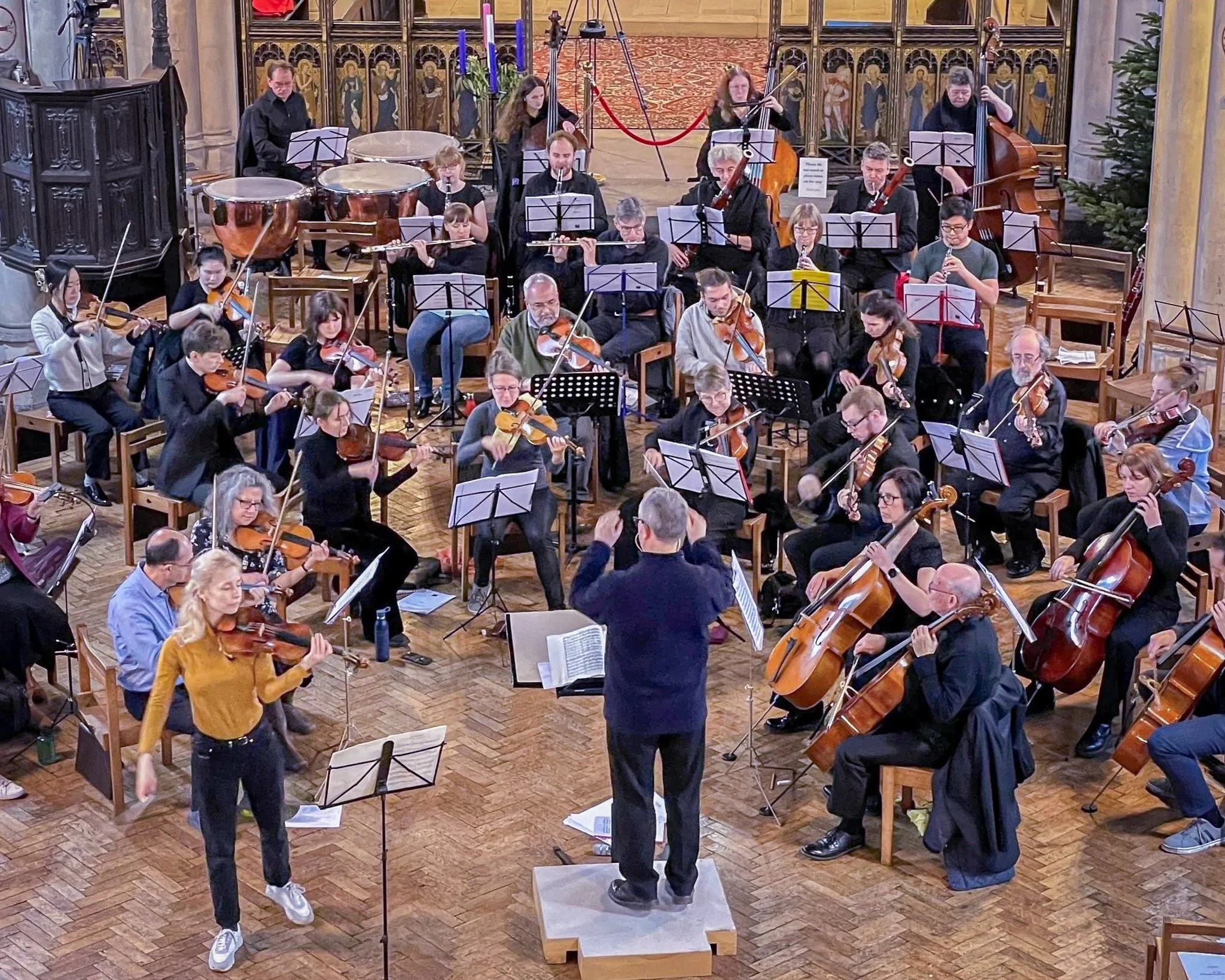 An orchestra performing in a church, conducted by a person standing on a small platform. The orchestra includes string, wind, and percussion instruments. The conductor is facing the orchestra with arms raised. The church interior features wooden floors, religious artwork, and a Christmas tree in the background.
