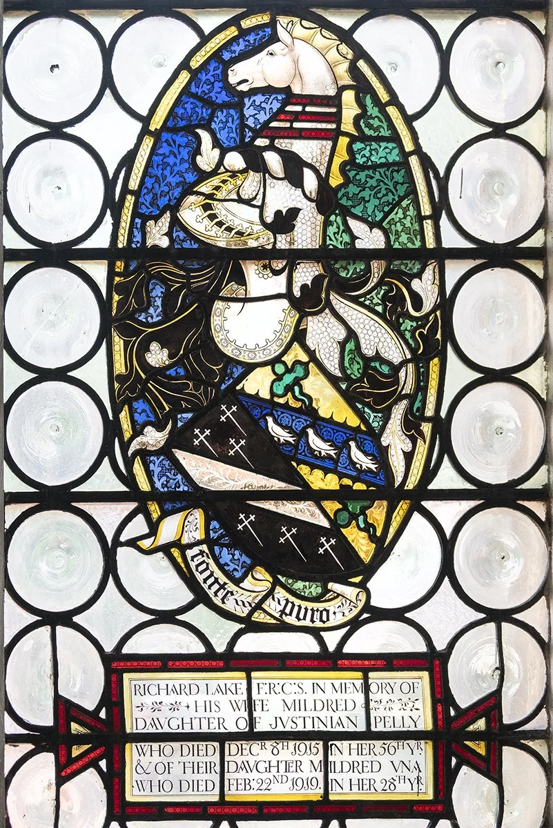 Stained glass window featuring a unicorn with a shield and banner with Latin-like inscription, surrounded by circles, with memorial text at the bottom honoring a woman named Mildred, daughter of Justinian Pelly, who died in 1919.