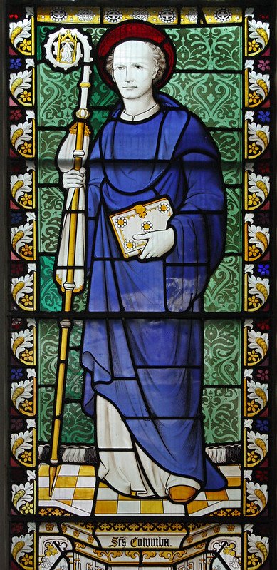 Stained glass window depicting a person in medieval attire holding a book, dressed in blue with a red hat and holding a staff, with intricate green and yellow background patterns.