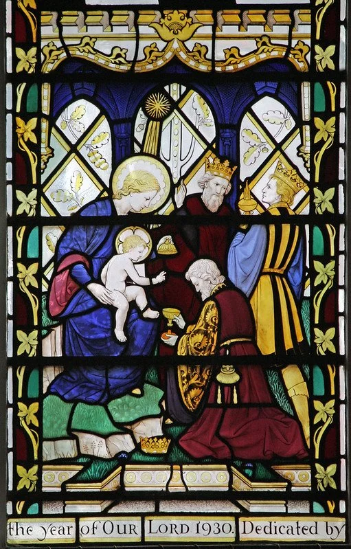 Stained glass window showing the nativity scene with Mary, Joseph, and baby Jesus, with a kneeling figure, likely a king or wise man, presenting a gift. The scene is decorated with gold accents and floral patterns, and includes the inscription "the y