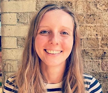 A woman with long blonde hair smiling, wearing a striped shirt against a brick wall background.